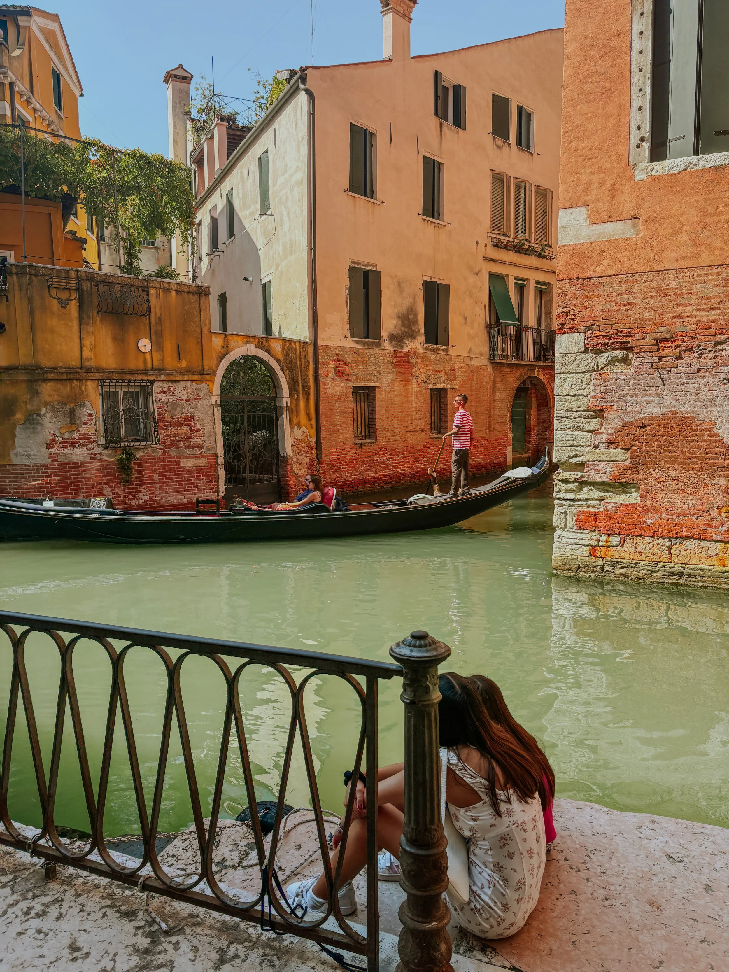 A girl sitting on the ground by a canal, with a metal railing in front of her, in Venice, Italy. A gondola with a gondolier is on the water, and old buildings line the canal.
