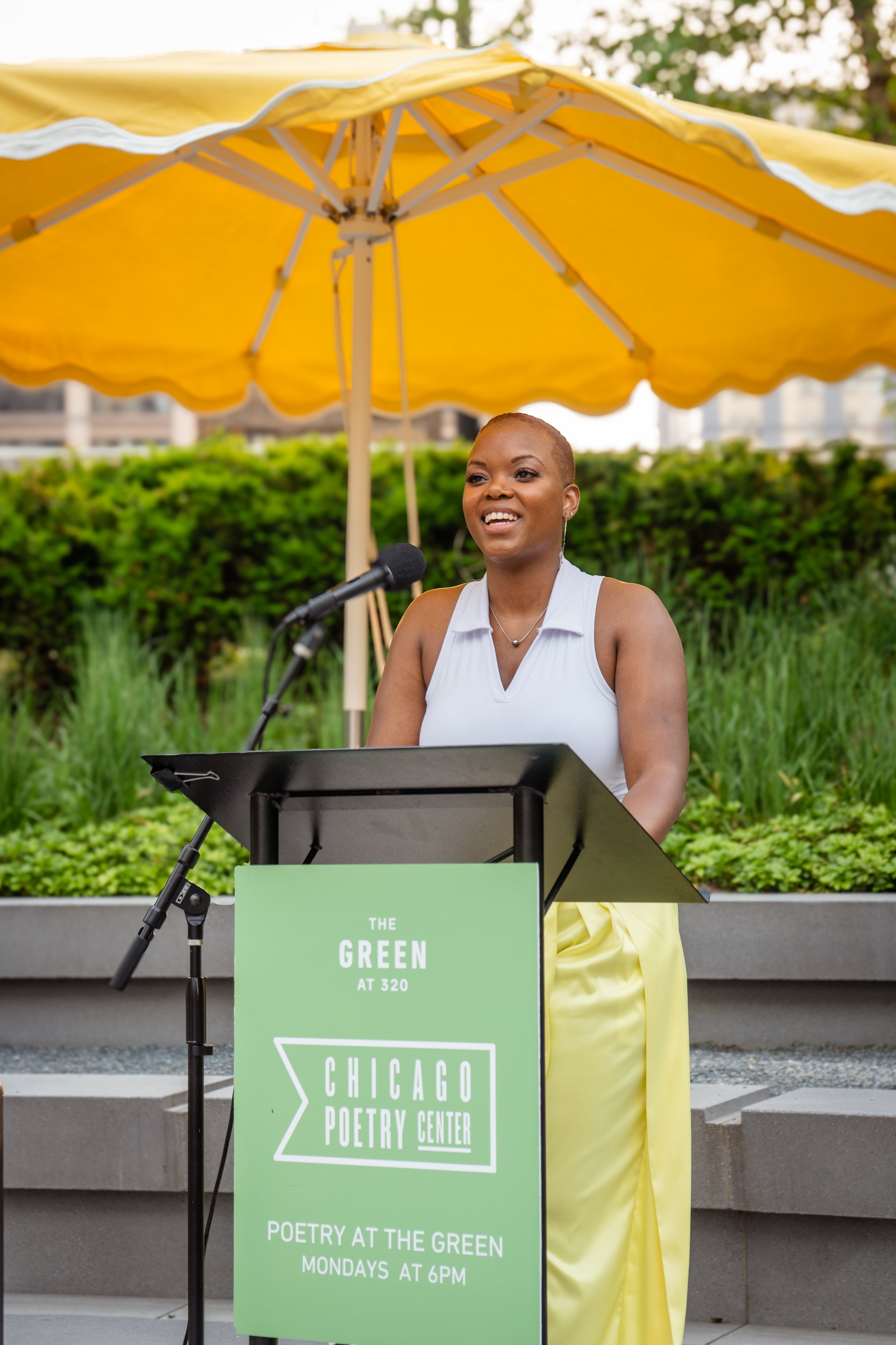 A woman standing at a podium under a yellow umbrella outdoors, speaking with a microphone, at the Chicago Poetry Center event.