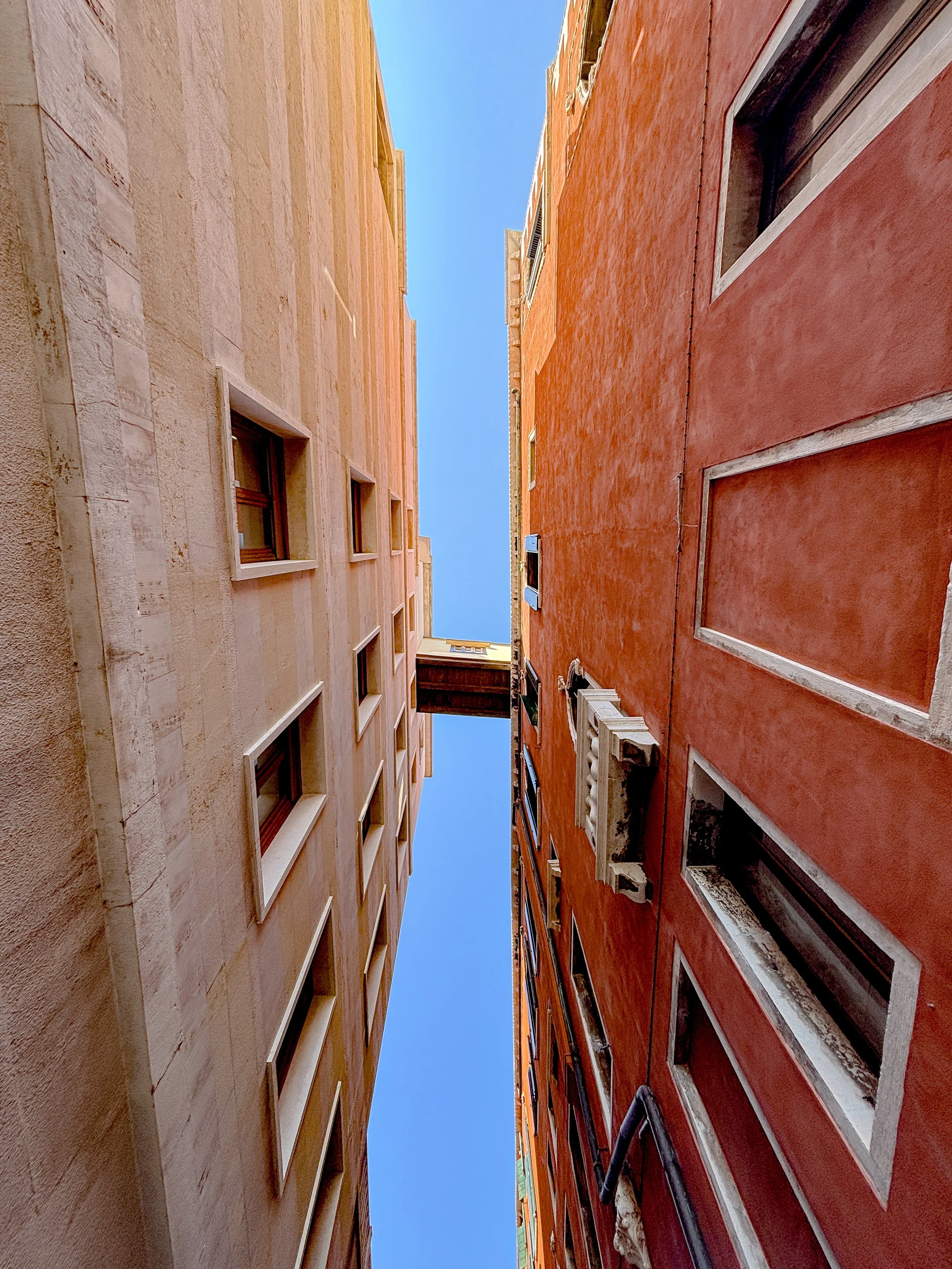 Low-angle view of two contrasting tall building facades with small windows, one light-colored and the other reddish, against a clear blue sky.
