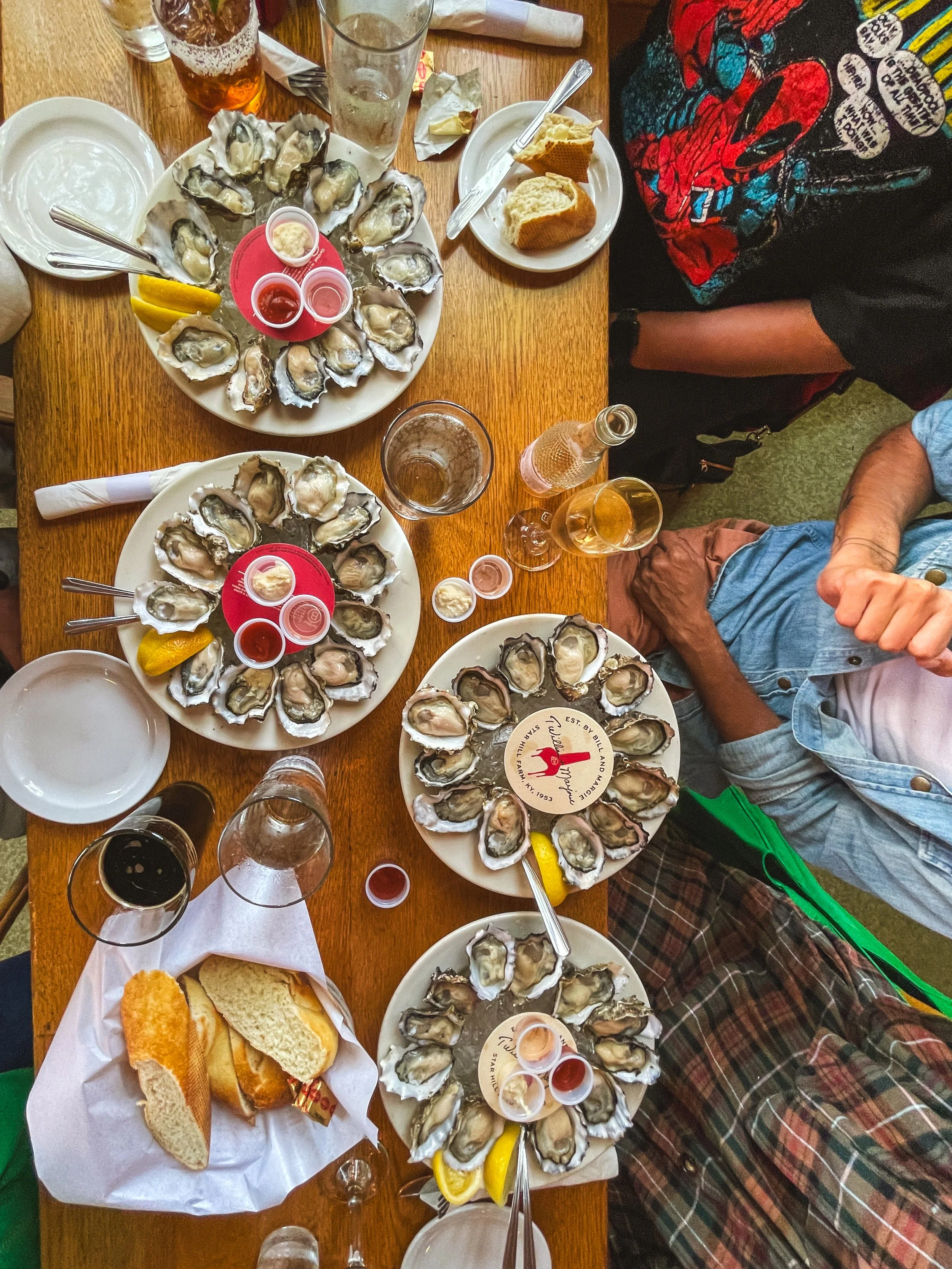 A table with several plates of oysters, some with lemon wedges, and small containers of hot sauce, along with glasses of drinks including beer and wine, and a plate with bread and garlic bread.