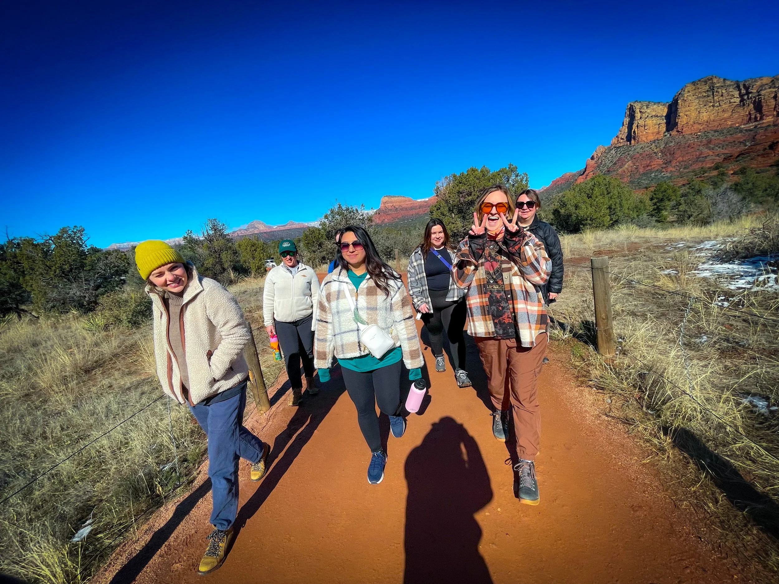 Group of six women hiking on a trail in a desert landscape with red rock formations in the background under a clear blue sky.