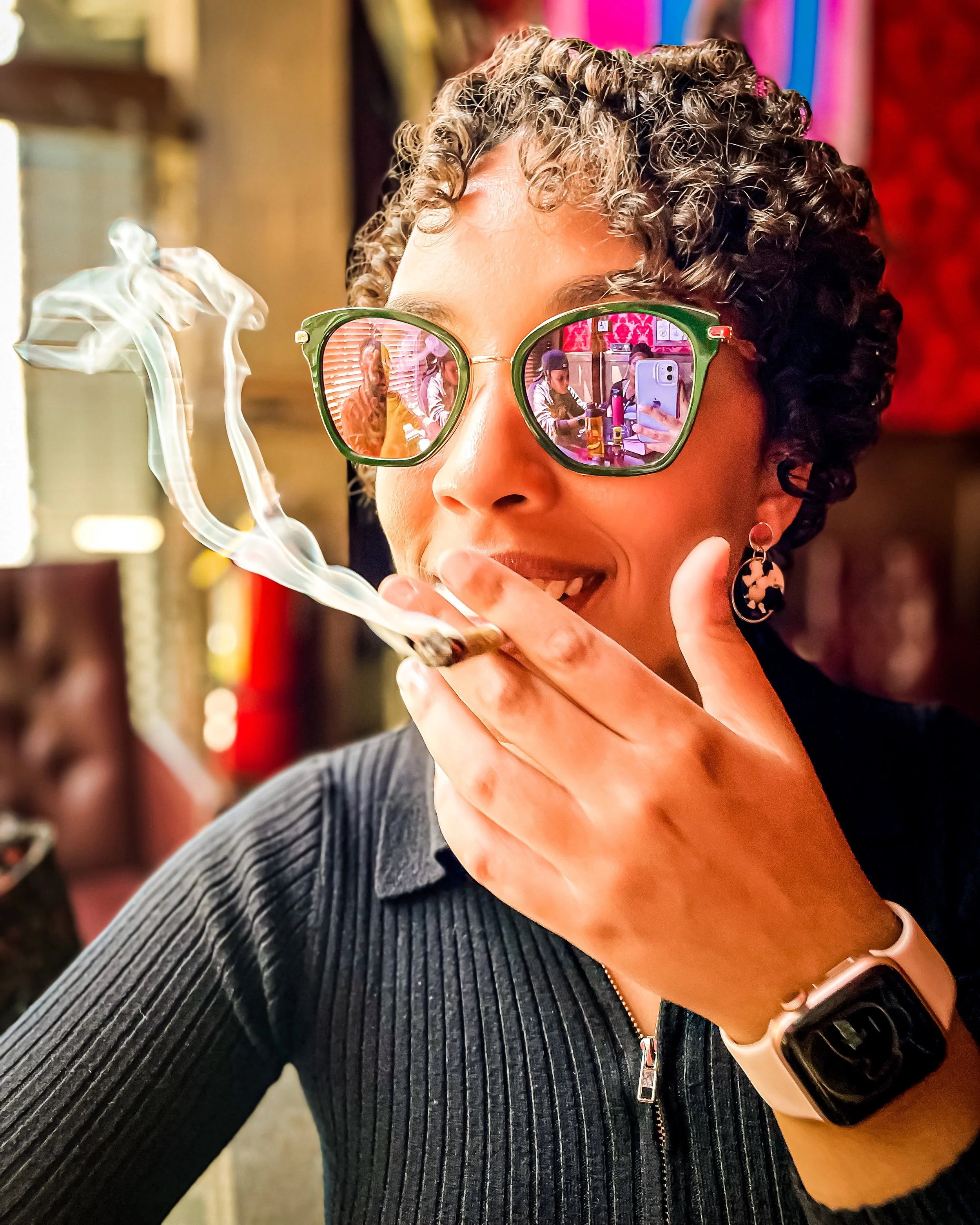 Woman with curly hair wearing green sunglasses, smoking a cigar, in a colorful, lively indoor setting.