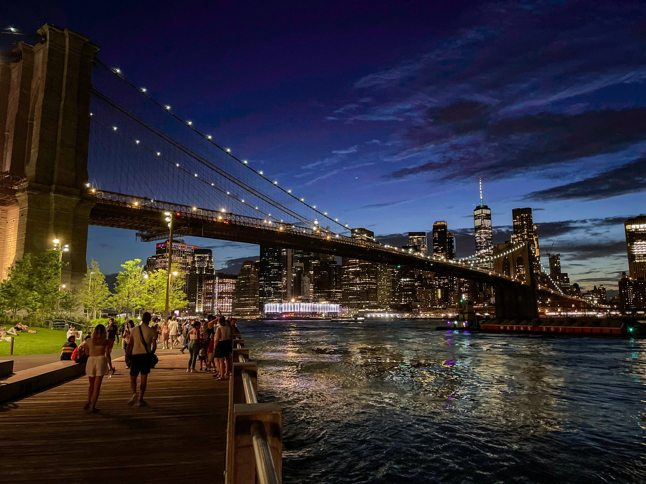 Nighttime view of the Brooklyn Bridge with Manhattan skyline and One World Trade Center lit up, people walking along the waterfront promenade.