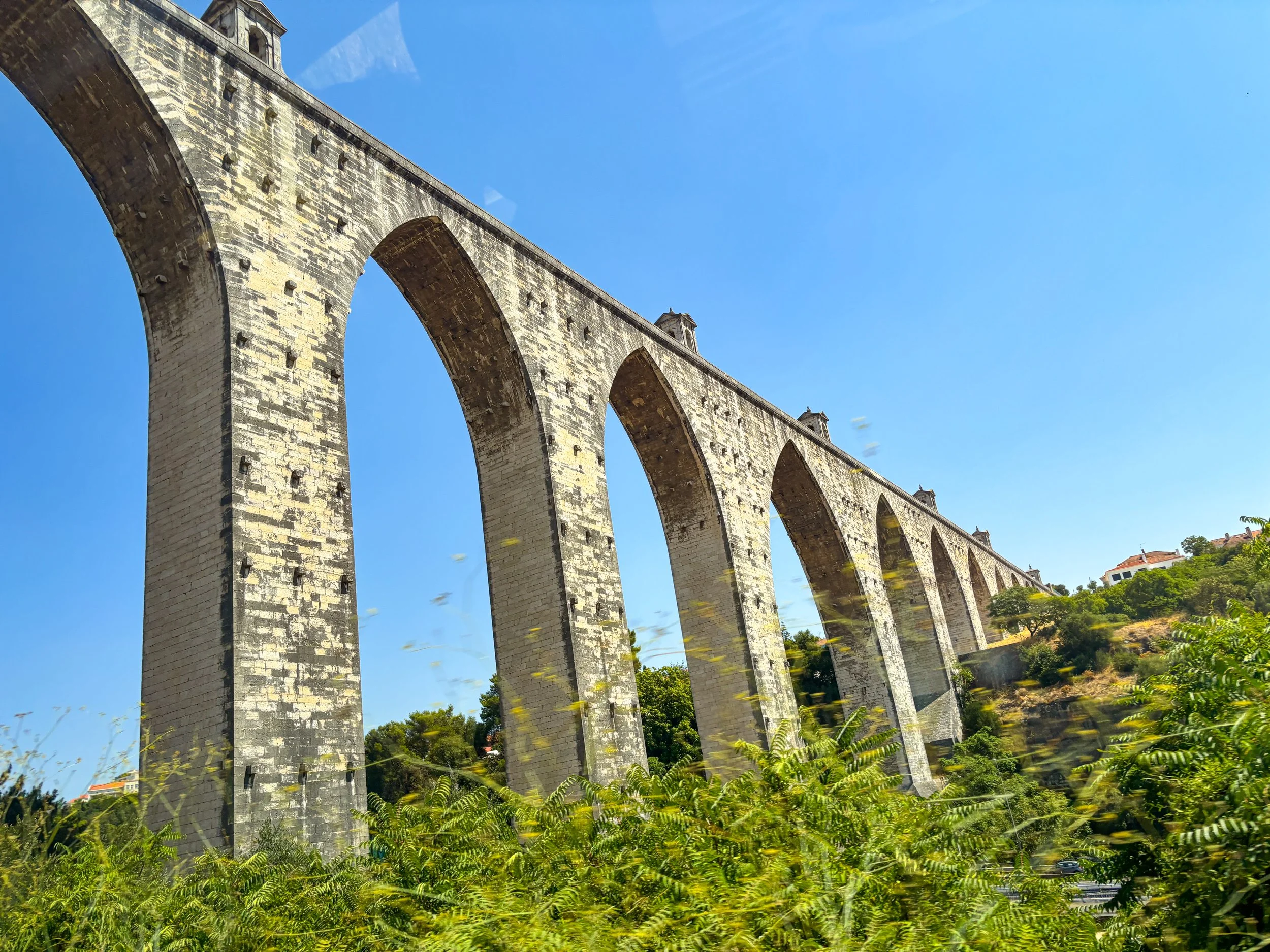 A large stone aqueduct with multiple arches extending into the distance, surrounded by green vegetation and under a bright blue sky.