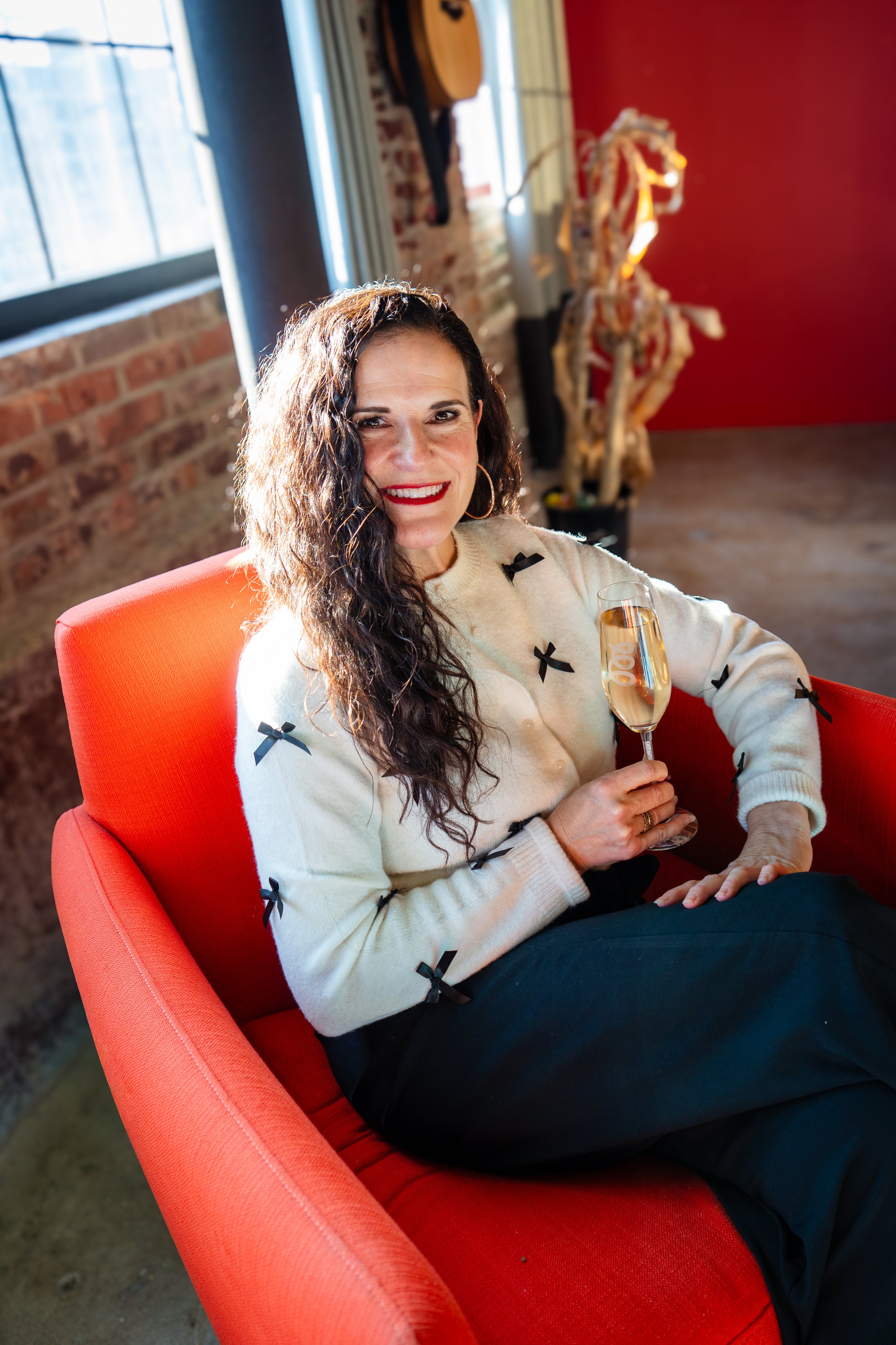 A woman with long, curly dark hair and red lipstick sitting on a red chair, holding a glass of champagne, smiling at the camera. There is a skeleton decoration and a guitar on the wall behind her, with a brick wall and window in the background.