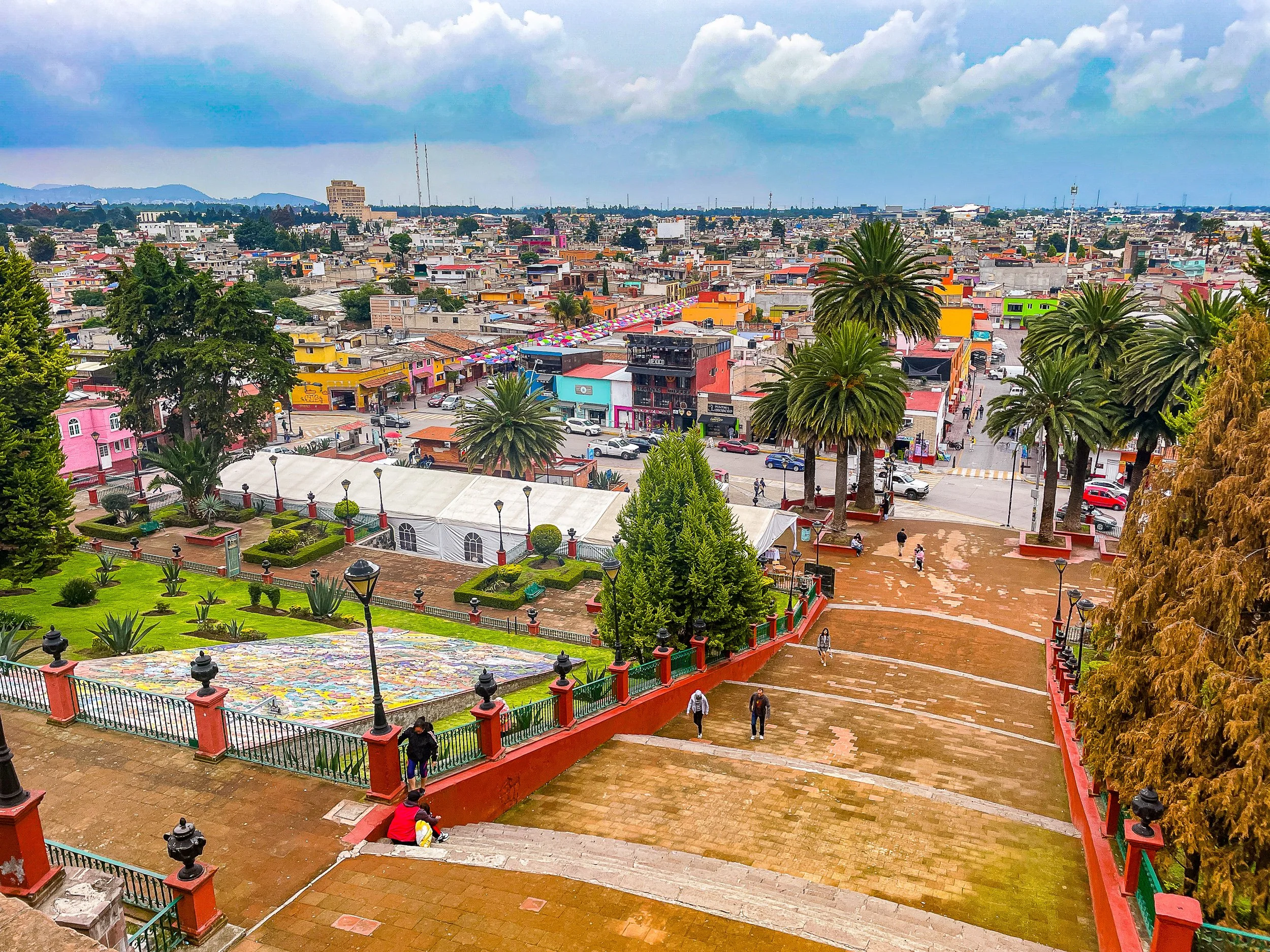 City view from a hilltop park with colorful buildings, palm trees, and people walking on paved pathways under cloudy sky.
