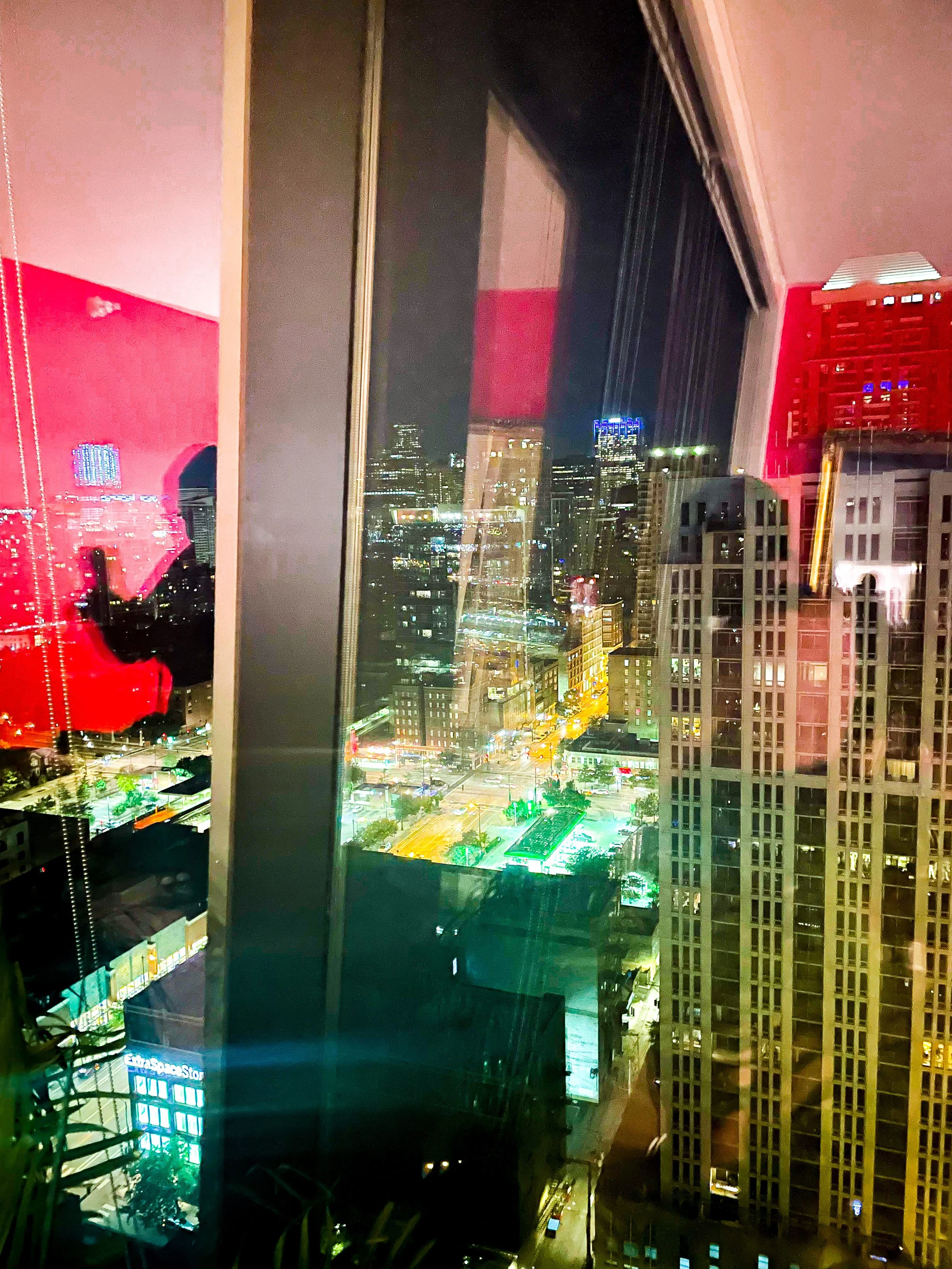 Night cityscape viewed through a window, reflecting indoor lights and city buildings.