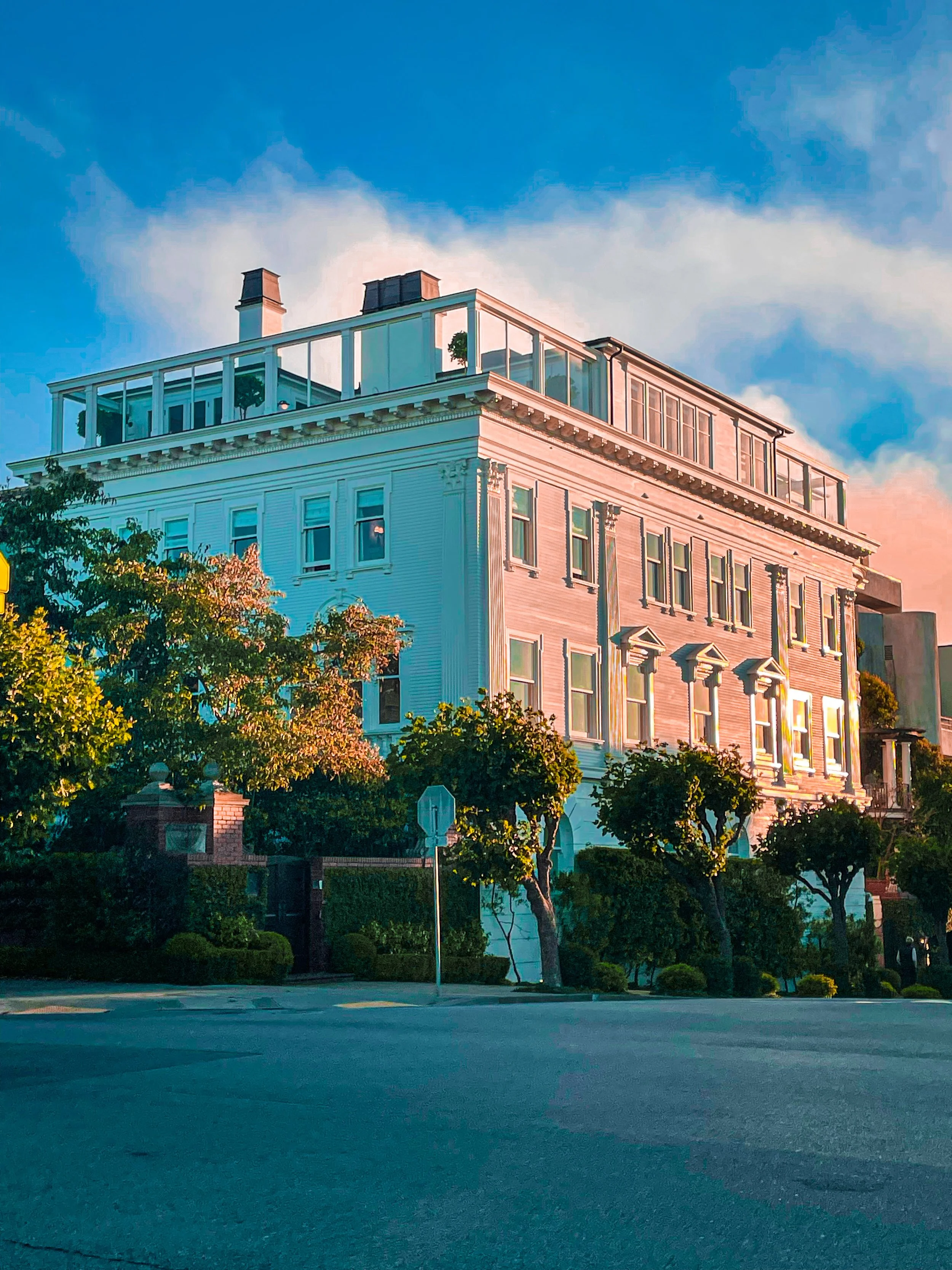A large, ornate residential building with pastel pink and white colors, multiple windows, decorative moldings, and a rooftop terrace with glass railing, surrounded by trees and a street in the foreground under a partly cloudy sky.