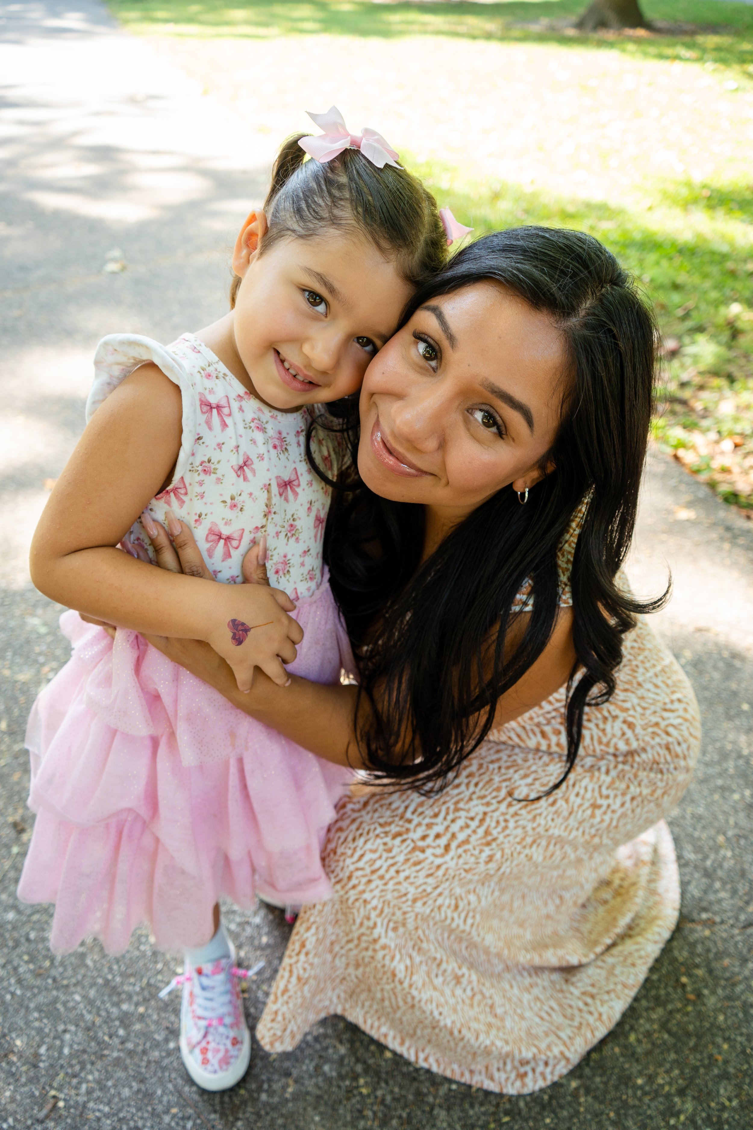 A woman and a young girl hugging outdoors on a sunny day, with trees and grass in the background.