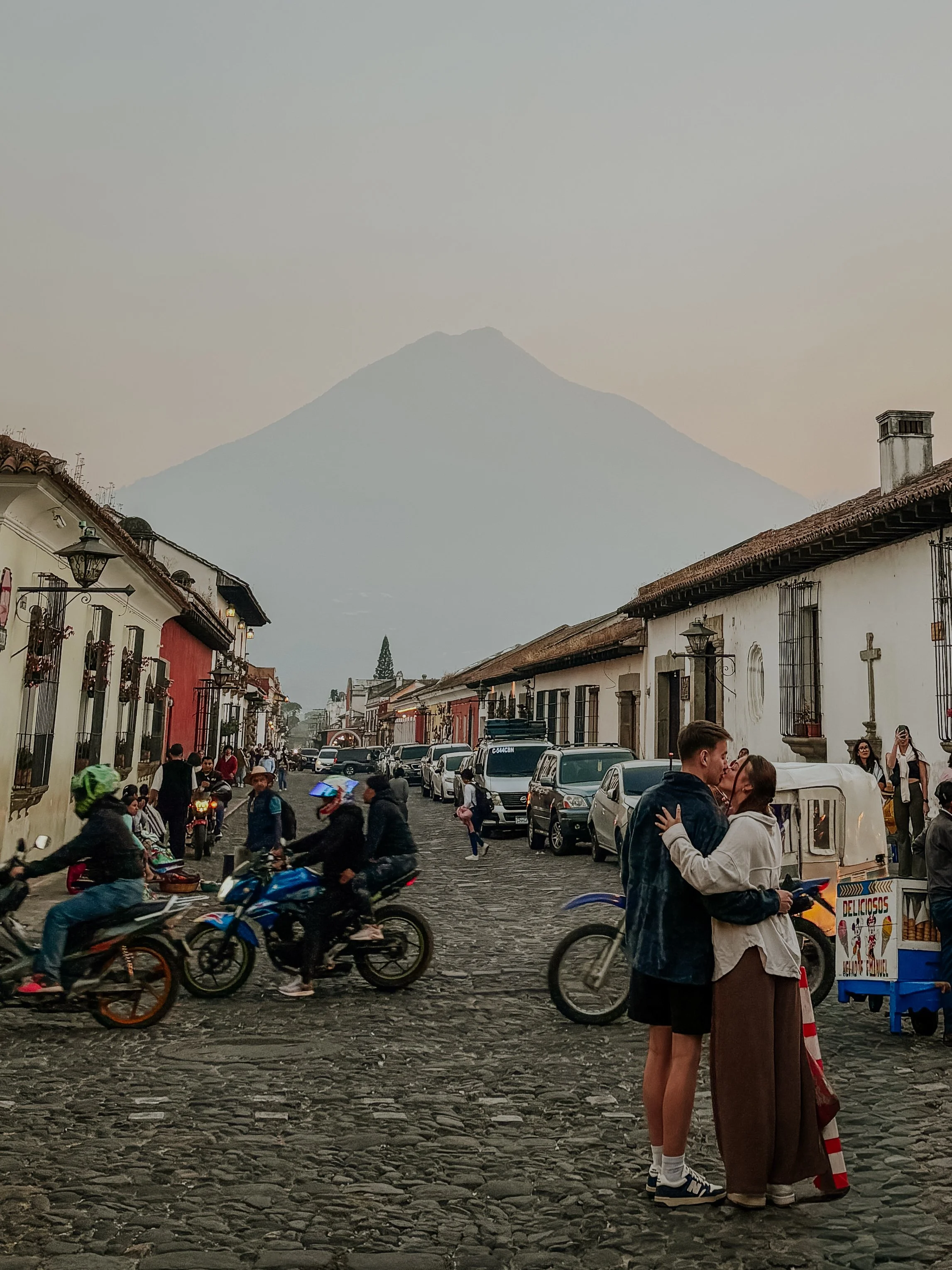 A lively street scene in a historic town with cobblestone streets, colorful colonial-style buildings, and a mountain in the background, with people on motorcycles, walking, and a couple kissing in the foreground.
