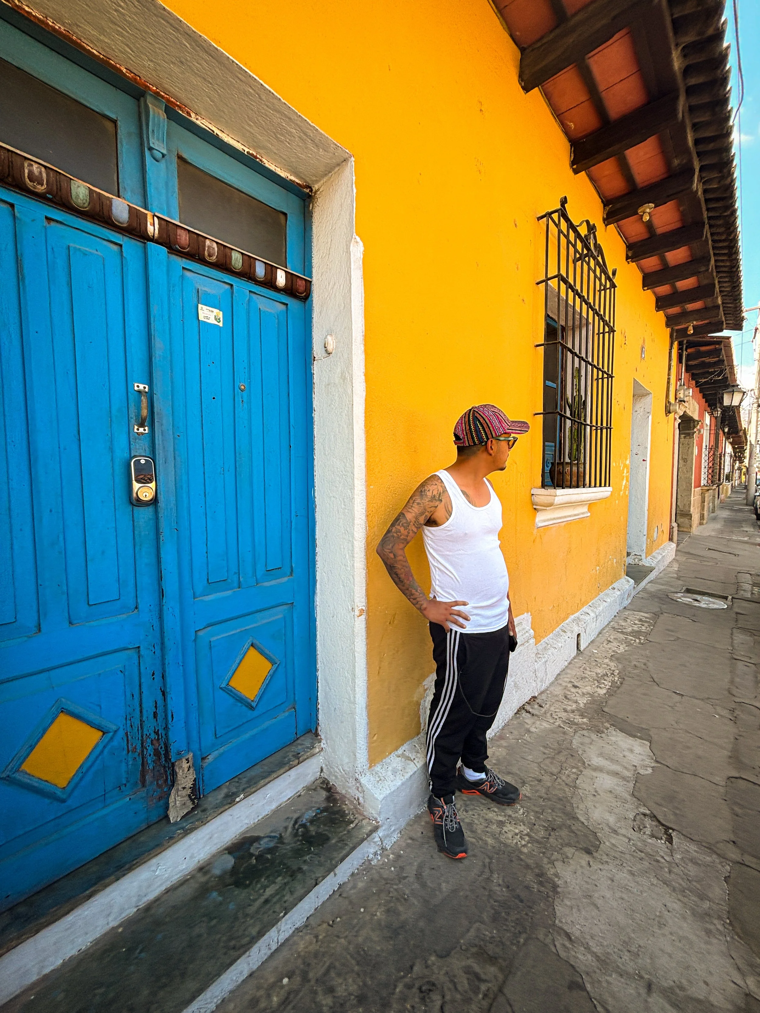 A man with tattoos wearing a white tank top, black pants with white stripes, black and orange sneakers, colorful hat, and sunglasses stands against a bright yellow wall with a blue door and barred windows on a sidewalk.