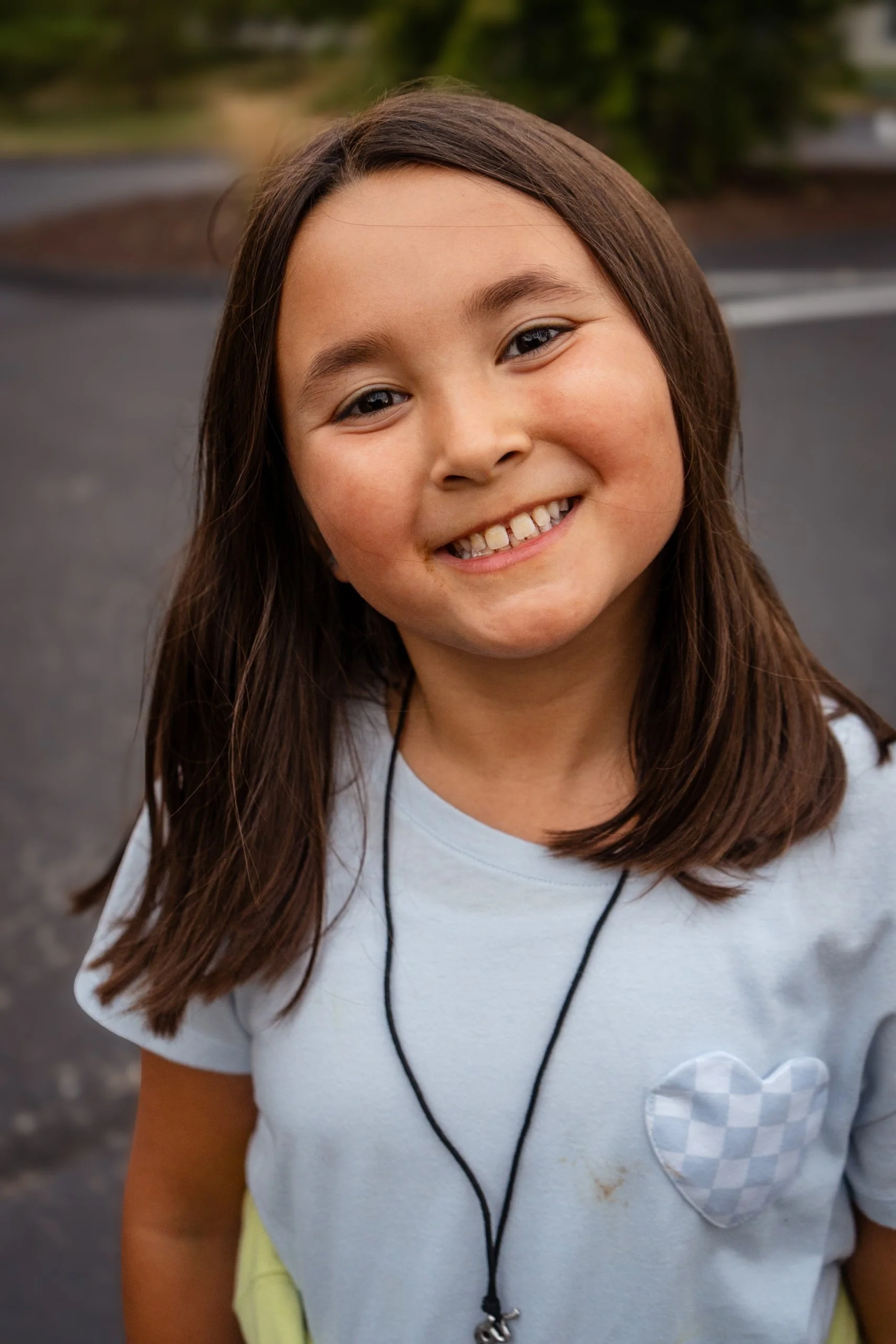 Close-up of a smiling young girl with brown hair wearing a light blue shirt with a checkered heart design on the chest and a necklace with a small cross pendant, standing outdoors on a paved street with greenery in the background.