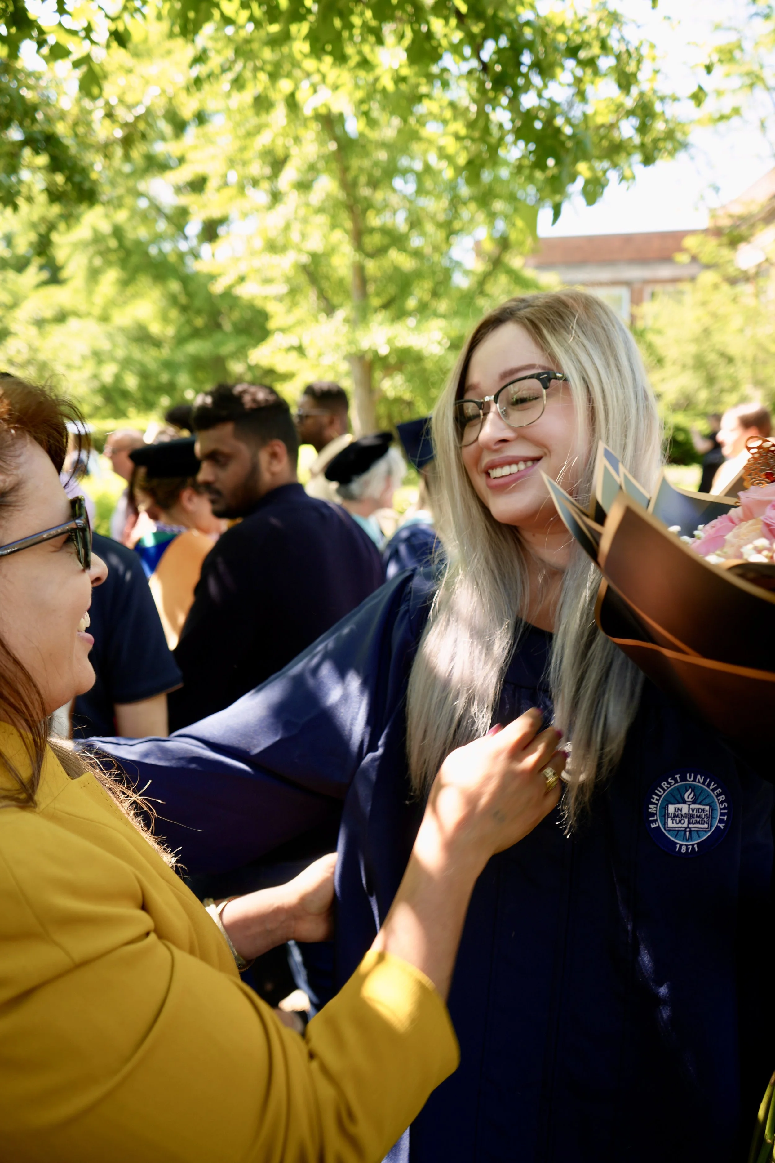 A young woman with blonde hair, glasses, and a Elmhurst University graduation gown smiling while receiving a flower bouquet from a woman in a yellow blazer during an outdoor graduation ceremony on a sunny day.