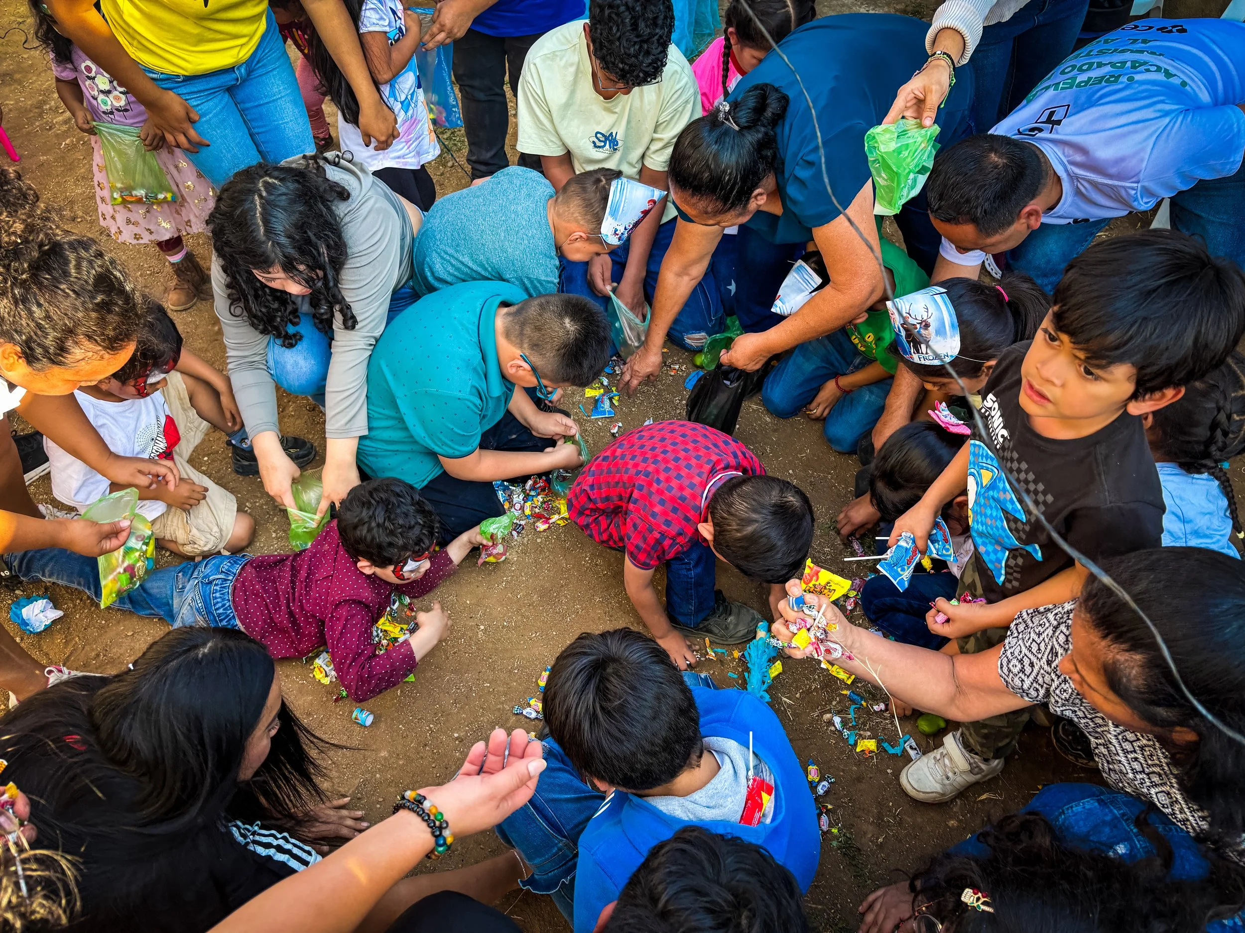 Group of children and adults gathered around on the ground, reaching for colorful candies and treats scattered on the dirt surface.