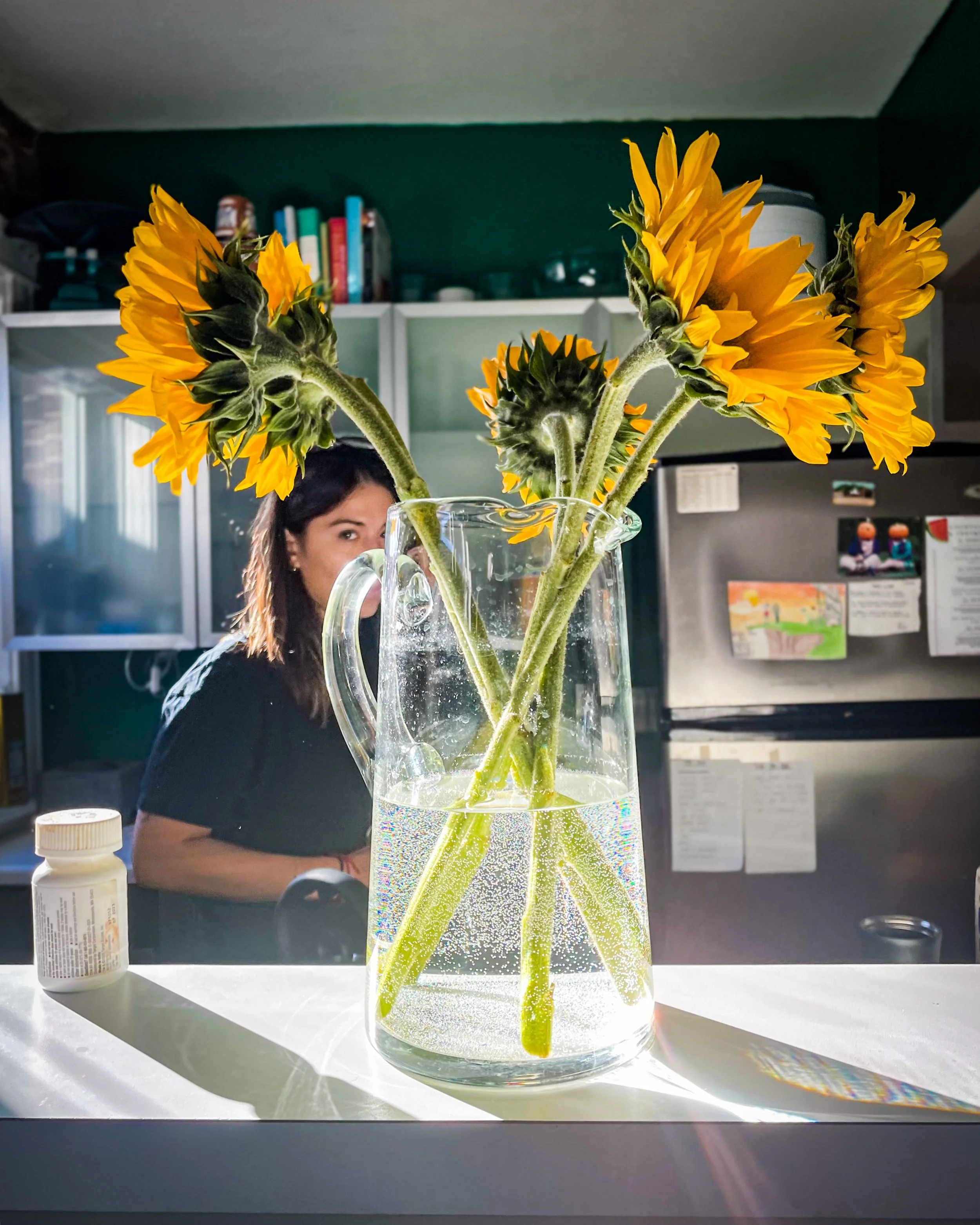 A clear glass pitcher with a handle contains three sunflowers in a kitchen setting, with sunlight creating shadows and rainbow reflections on the countertop.