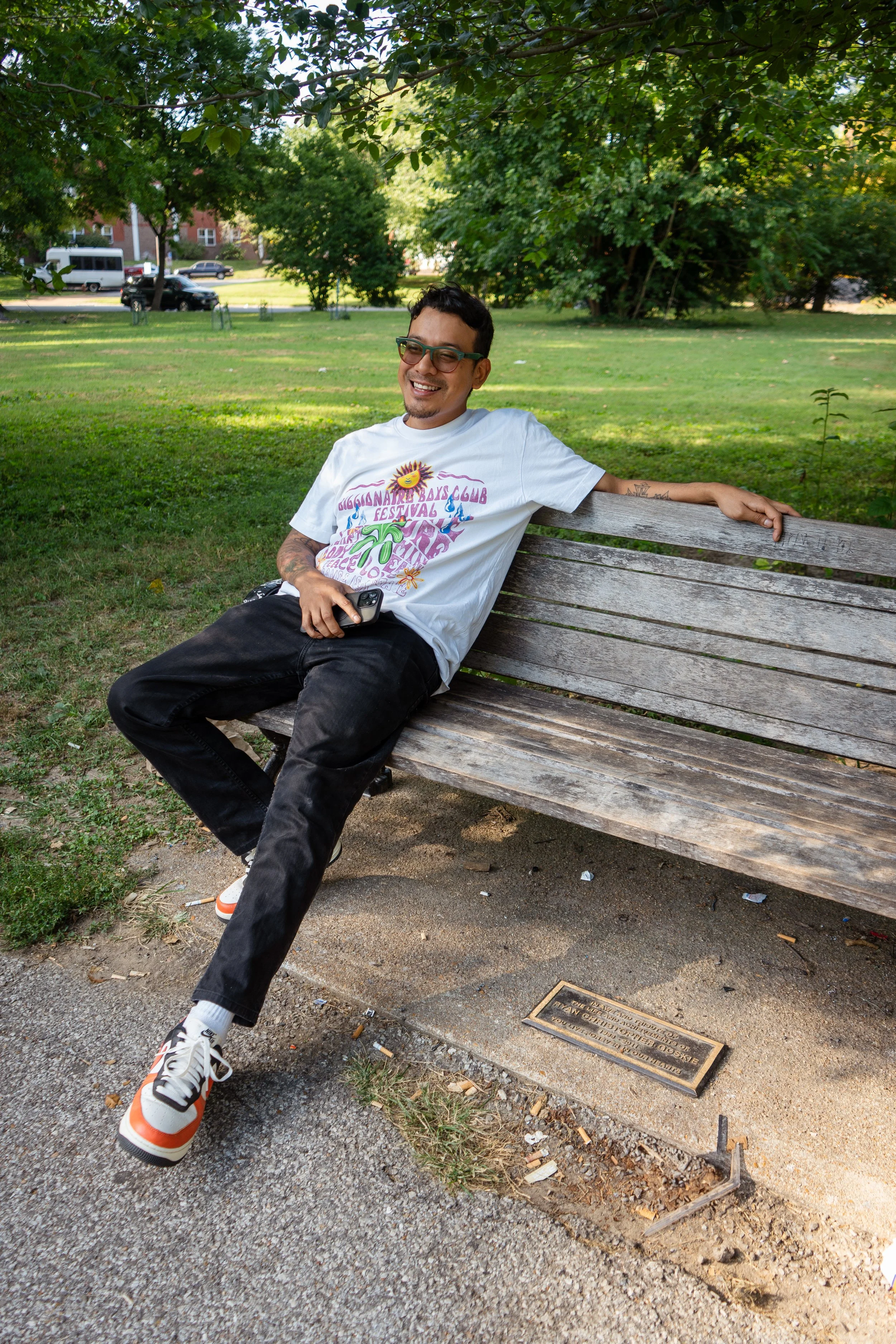 A young man sitting on a park bench, smiling and holding a phone, wearing sunglasses and a white T-shirt with colorful print, with lush green trees and grass in the background.