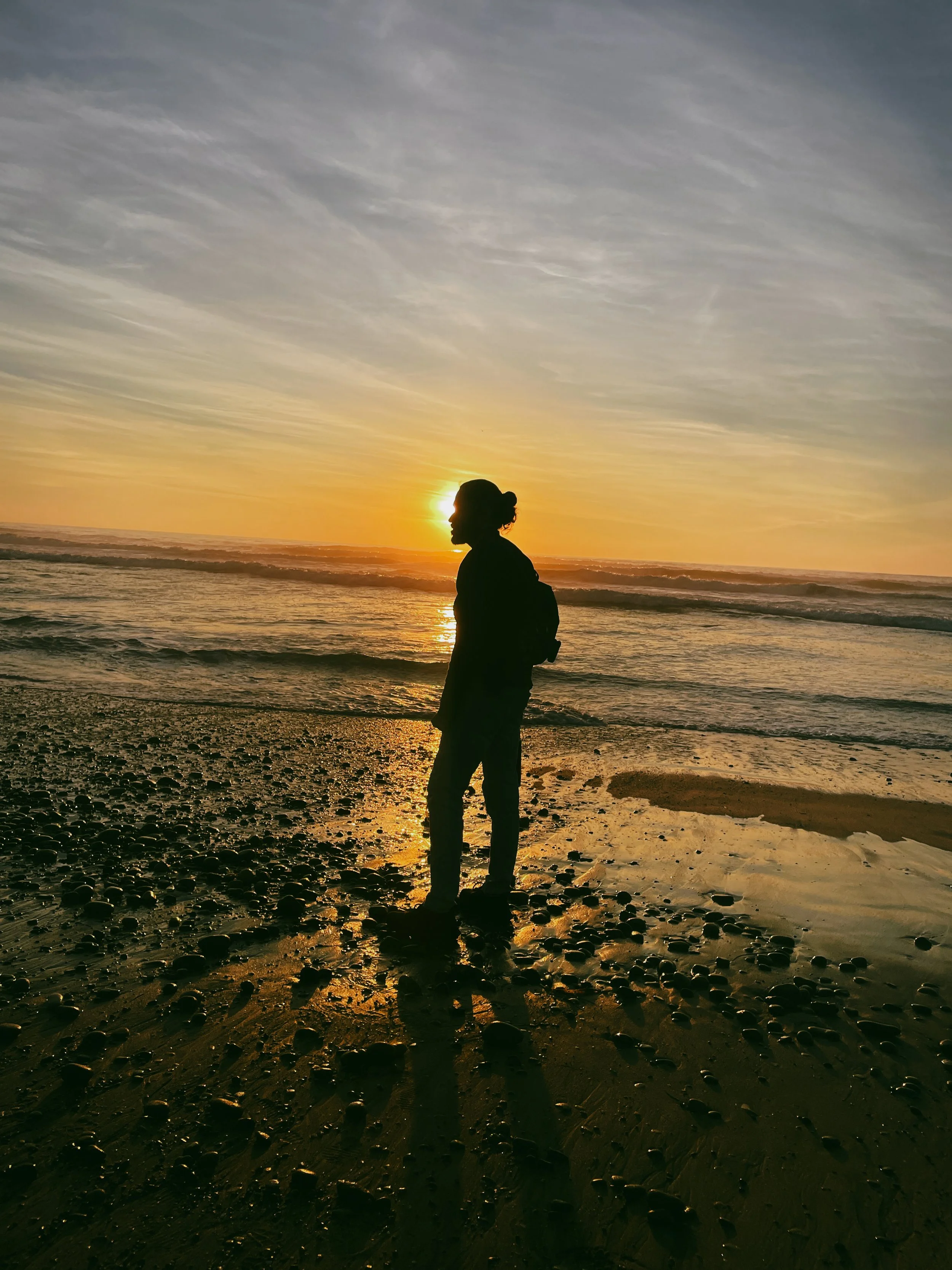 Silhouette of a person standing on a rocky beach at sunset, with the sun low on the horizon and reflecting on the water.