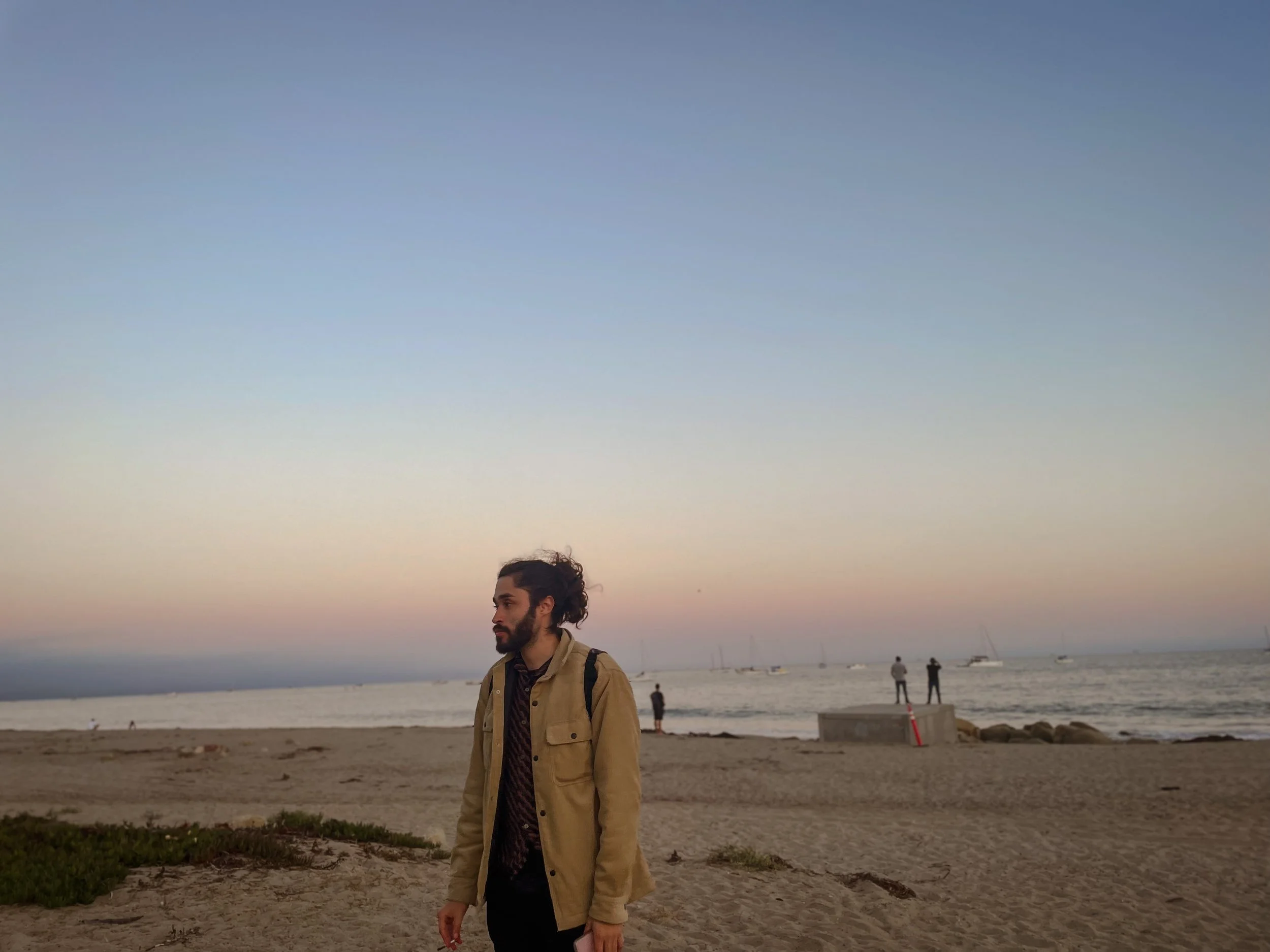 A man with a beard and long curly hair tied back, wearing a tan jacket and backpack, walking on the beach at sunset, with a calm ocean and sailboats in the background.