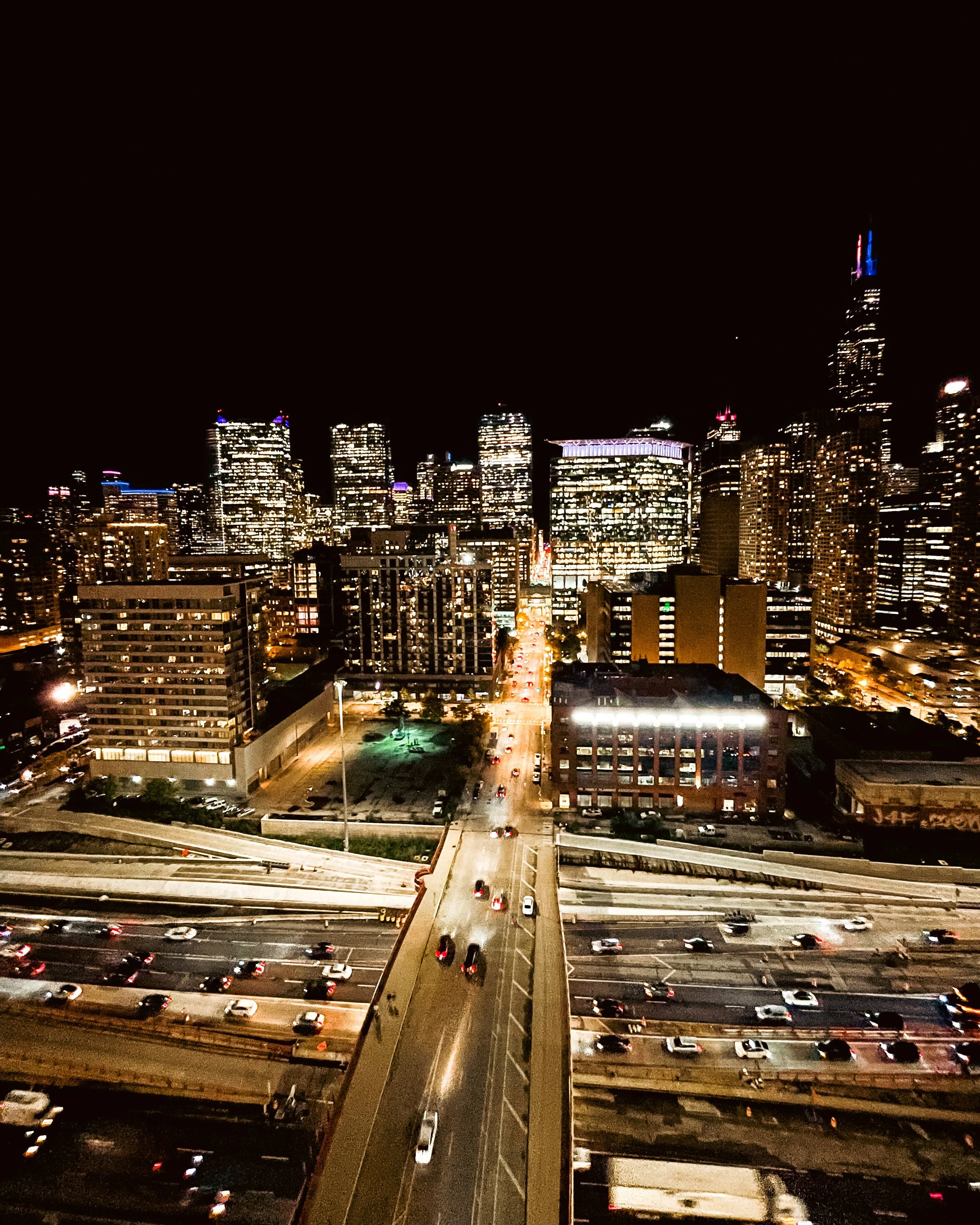 Nighttime view of a city skyline with illuminated skyscrapers and buildings, busy roads with car headlights, and a dark sky.