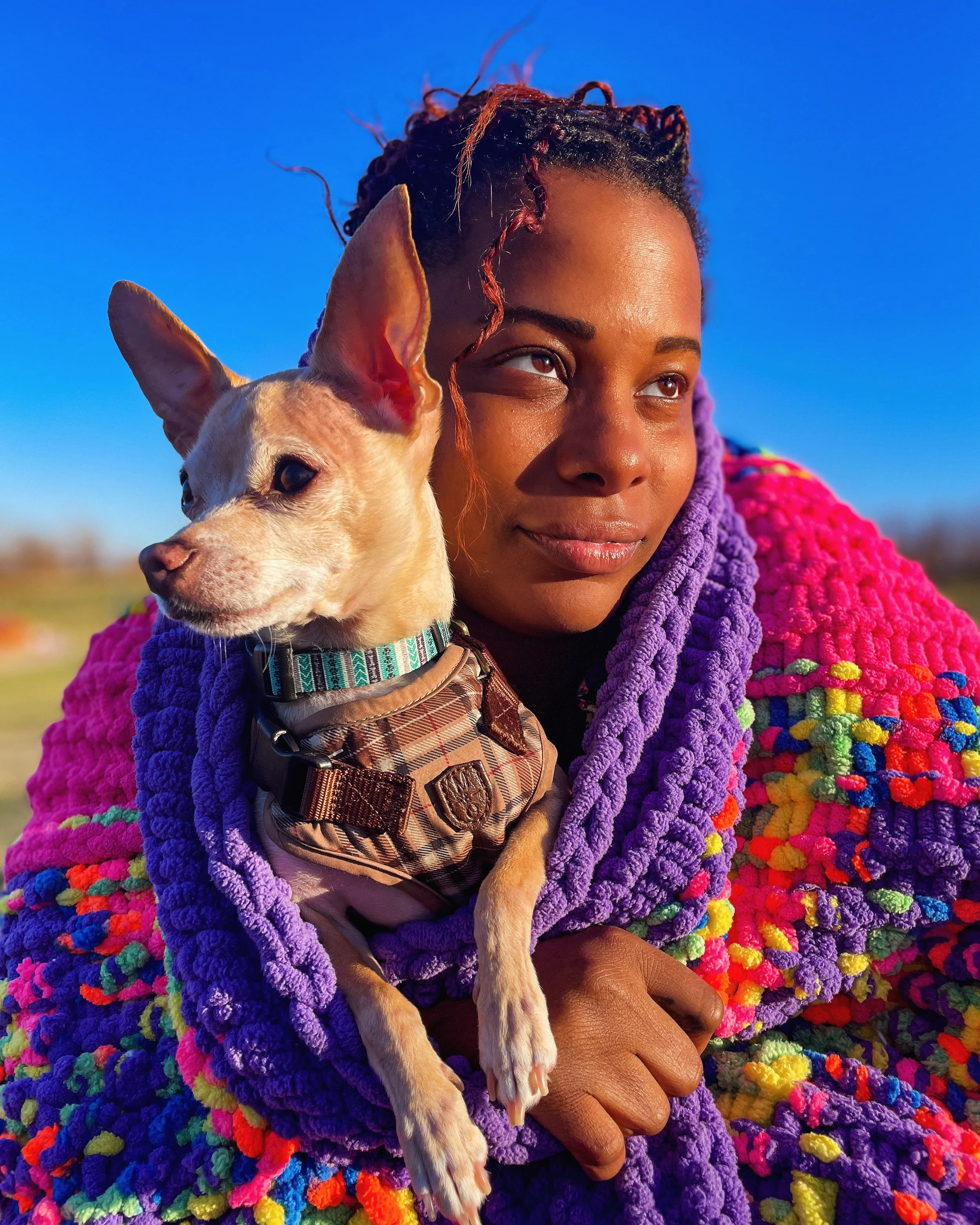 A woman outdoors holding a small dog wrapped in a colorful, textured blanket during sunset.