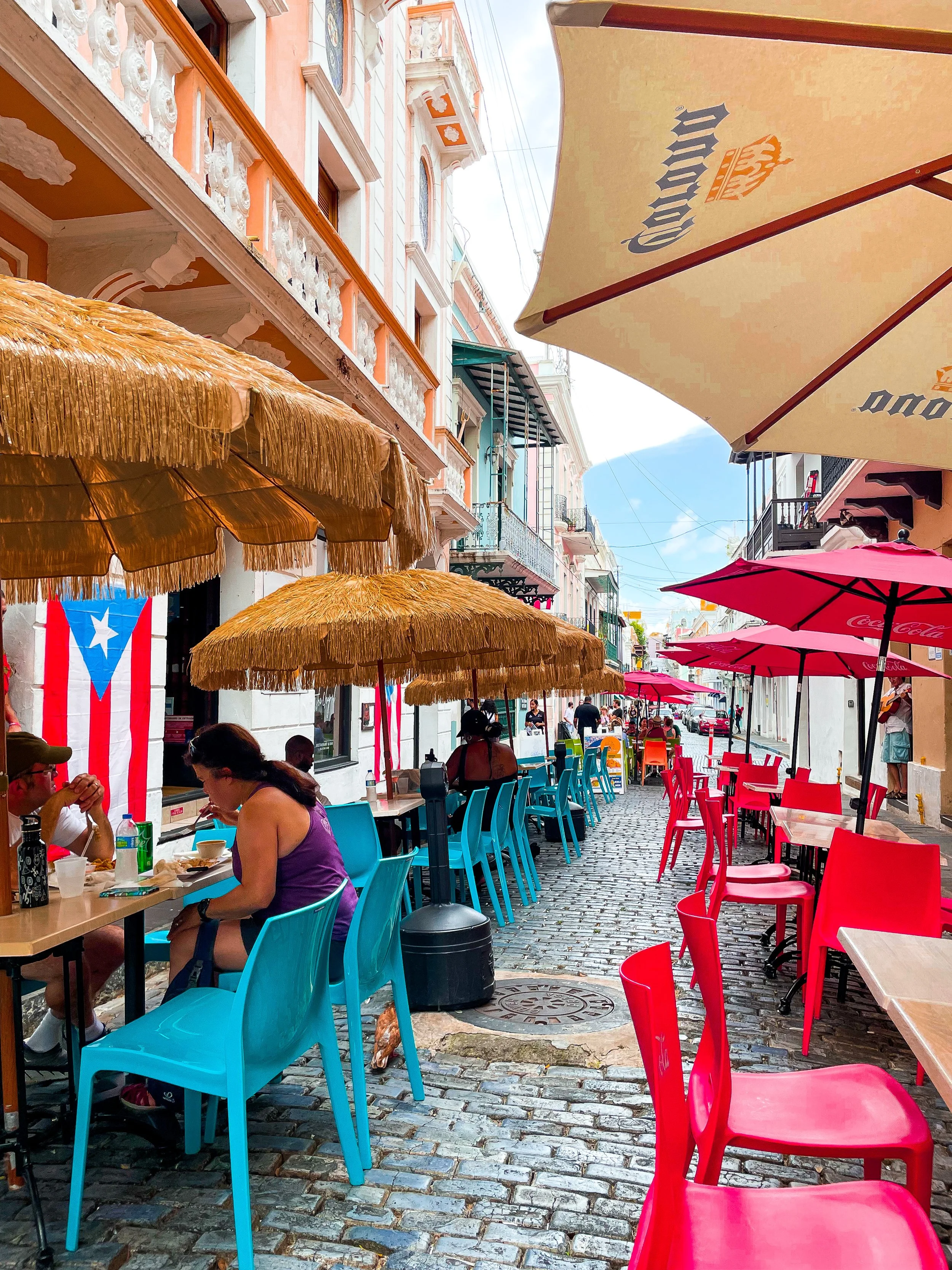 Colorful outdoor cafe seating with red, blue, and green chairs, thatched umbrellas, and a paved street with historic buildings and a Puerto Rican flag in the background.
