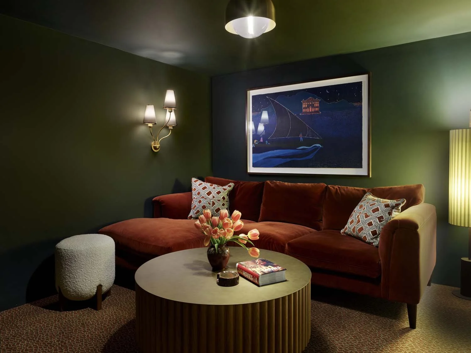 Living room with a velvet sofa, wall sconces, a painting, a round coffee table with flowers, books, and an ottoman, with a yellow floor lamp.
