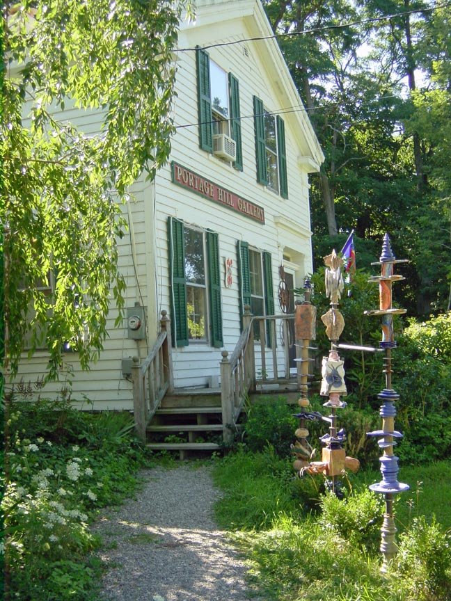 A white, two-story house with green shutters, surrounded by trees and greenery, with a wooden staircase leading to the entrance. There are colorful totem pole sculptures on the right side of the path leading to the house. The house has a sign that reads 'Portage Hill Gallery'.