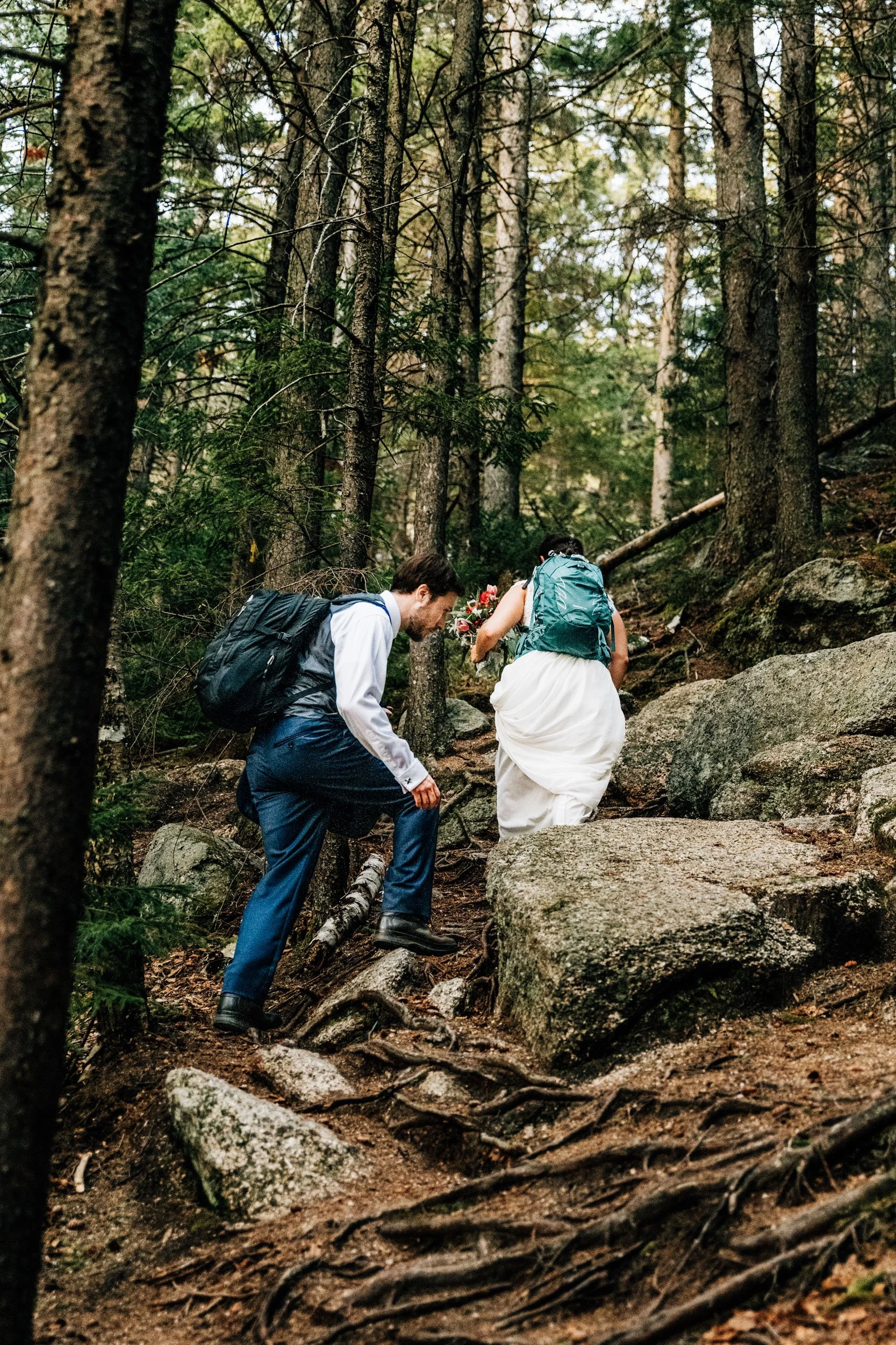 Bride and groom hiking a rooty forest trail with backpacks during a New Hampshire adventure elopement