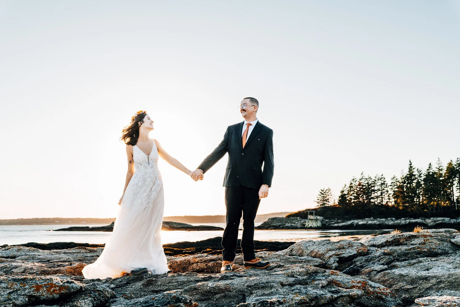 Alette and Kieron holding hands on the rocky shore of Pratts Island with the Maine coast behind them