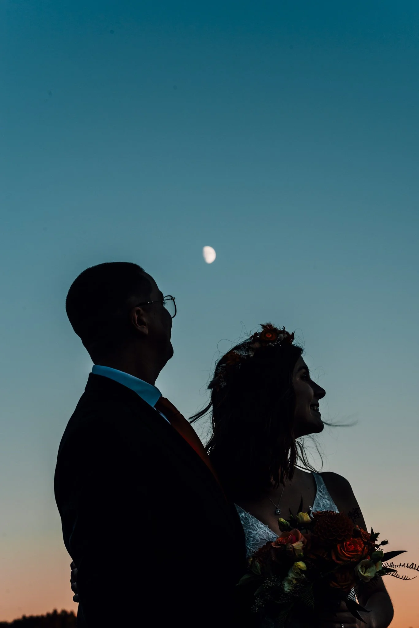 Silhouette of Alette and Kieron against a teal and amber dusk sky with the moon rising above them
