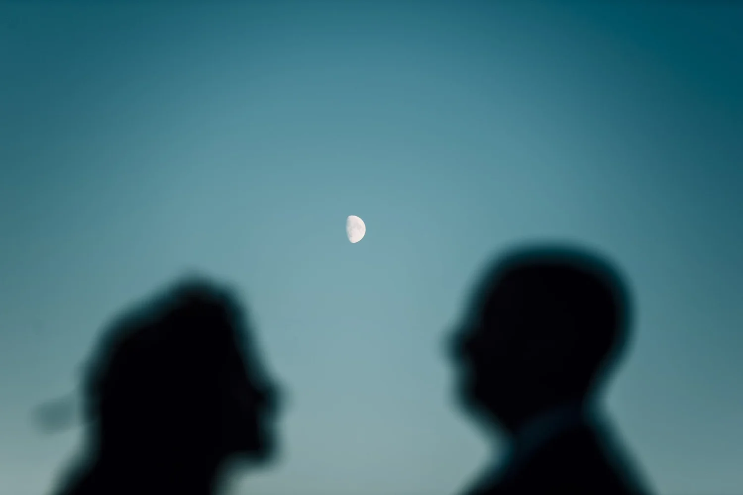 Blurred silhouettes of Alette and Kieron facing each other with the moon between them at blue hour
