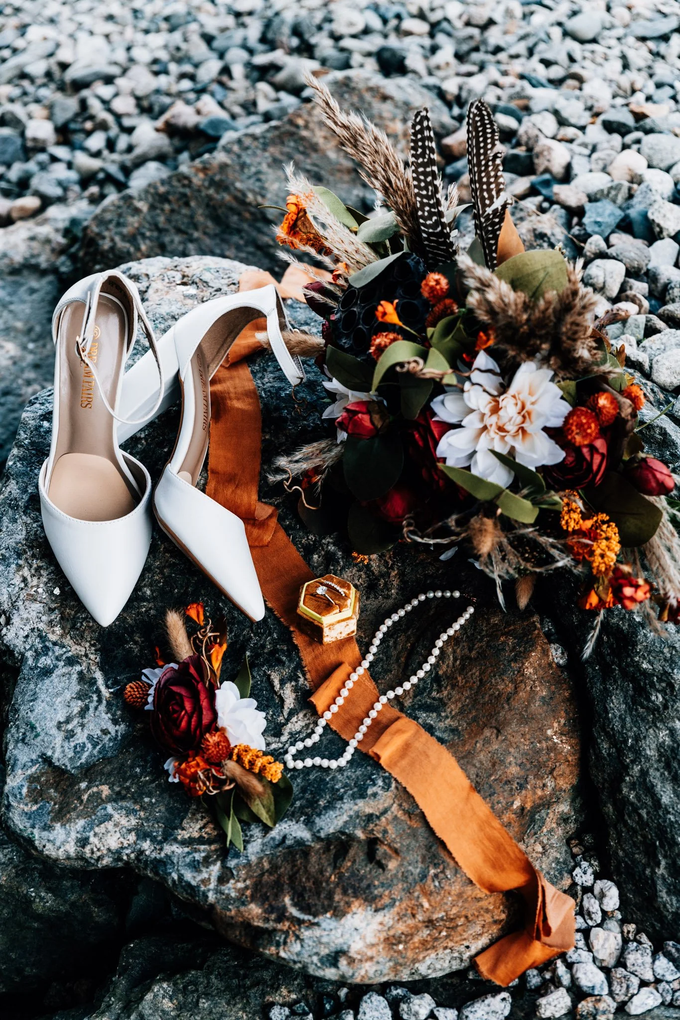 Wedding day details on Mount Washington granite: white heels, a rust and burgundy bouquet with feathers, a gold ring box and a pearl necklace