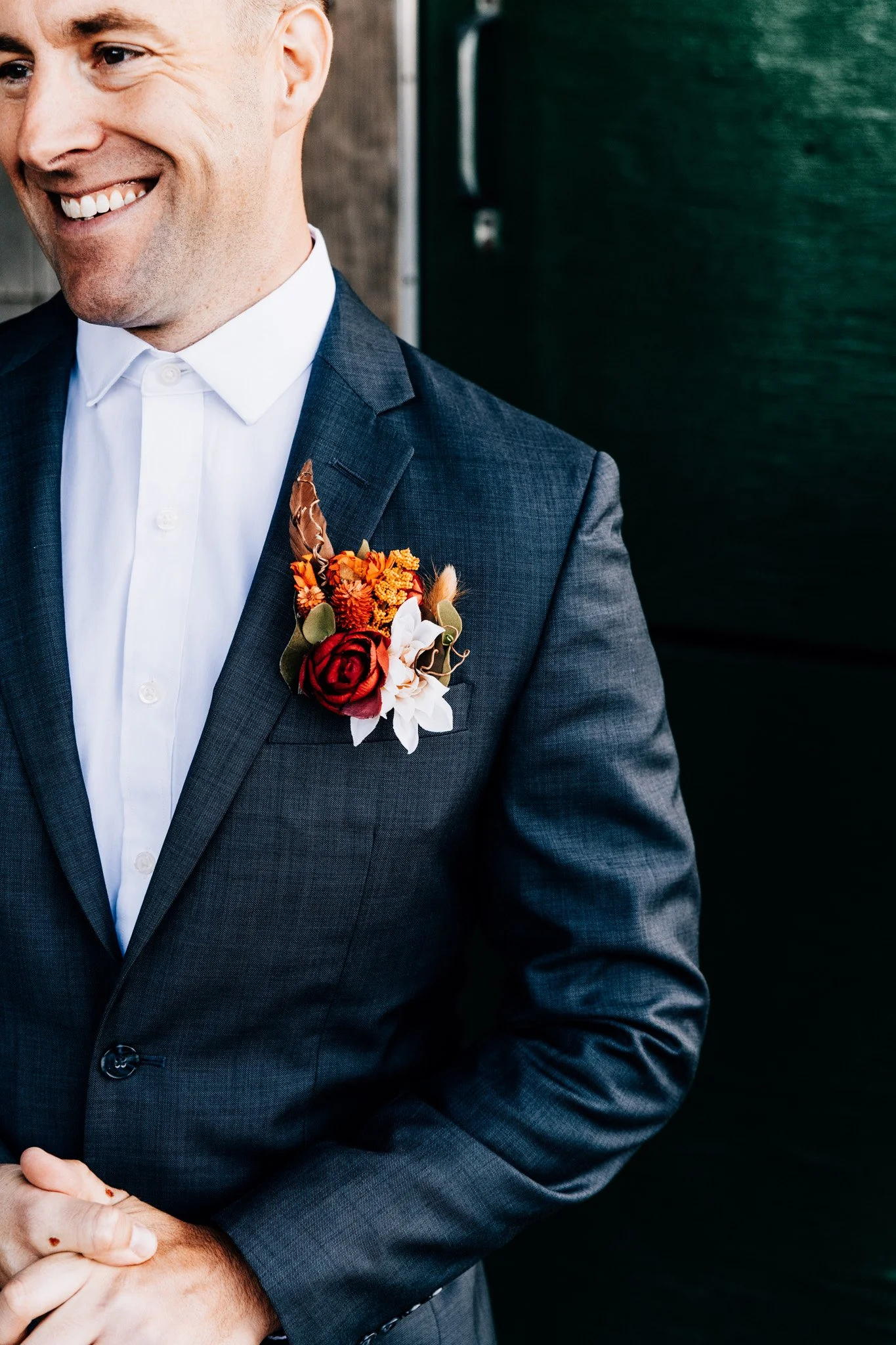 Groom smiling in a navy suit with a dried autumn boutonniere of roses, marigold and feathers