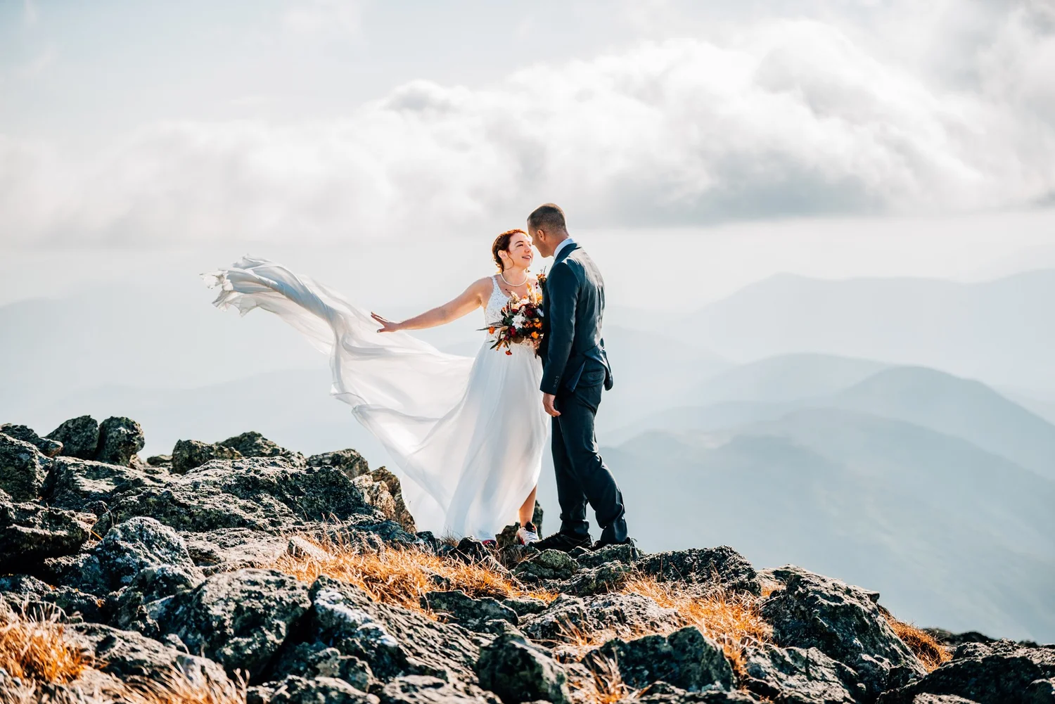 Wedding dress flying in the summit wind as Danielle and Tom lean in to kiss on Mount Washington