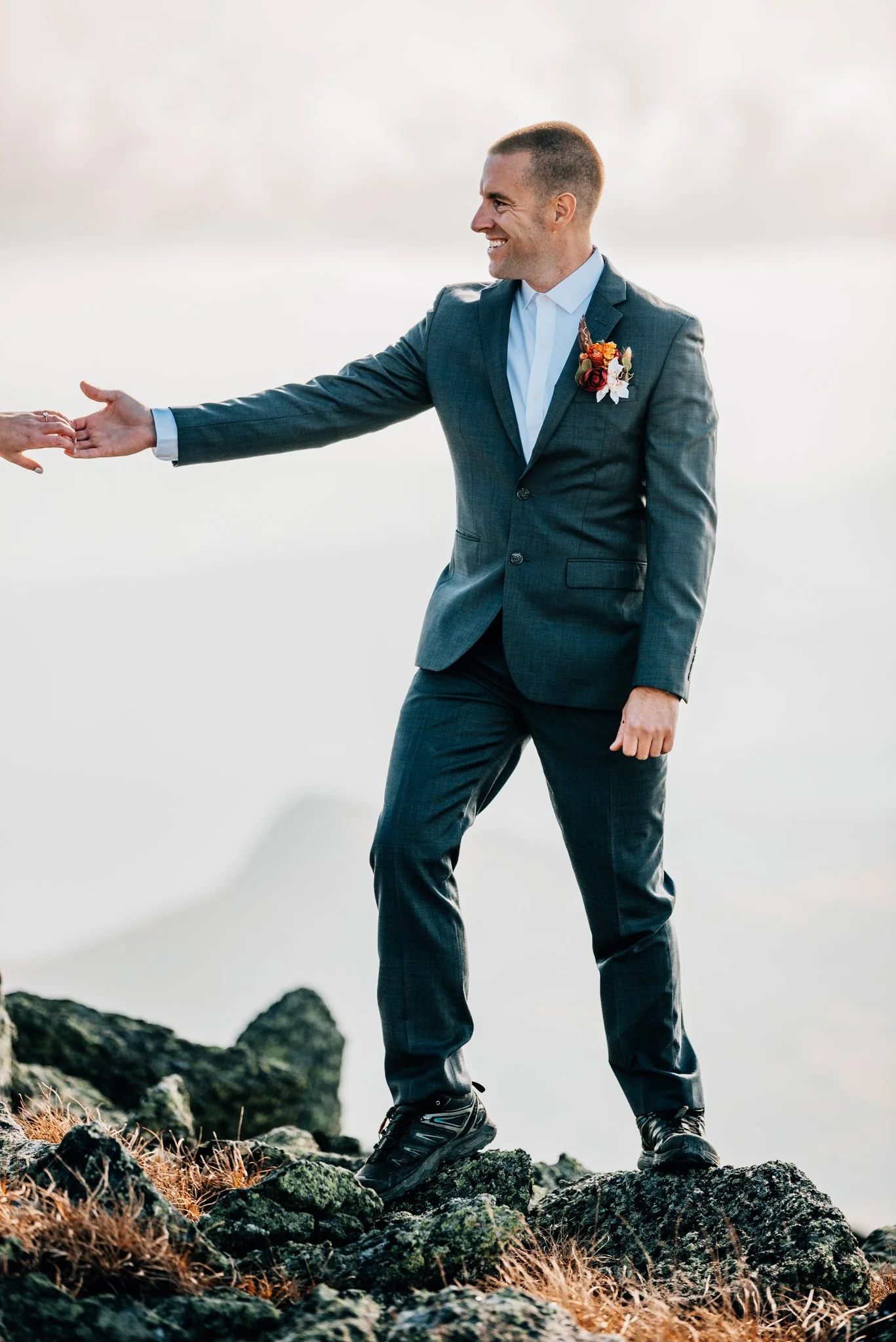 Groom in a charcoal suit reaching back for his bride's hand across the Mount Washington summit rocks