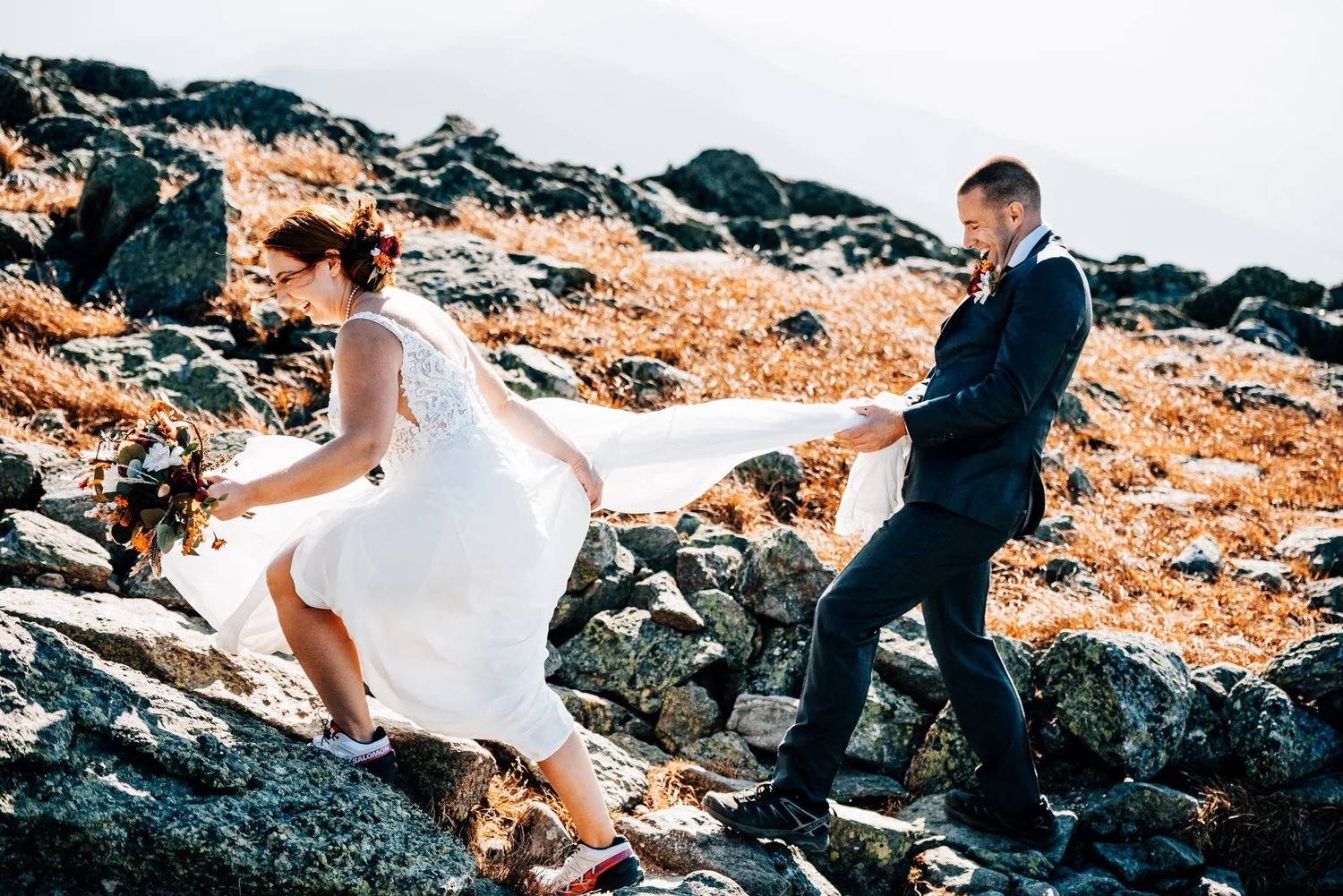 Groom carrying his bride's train as they scramble over rocks through the golden alpine grass of Mount Washington