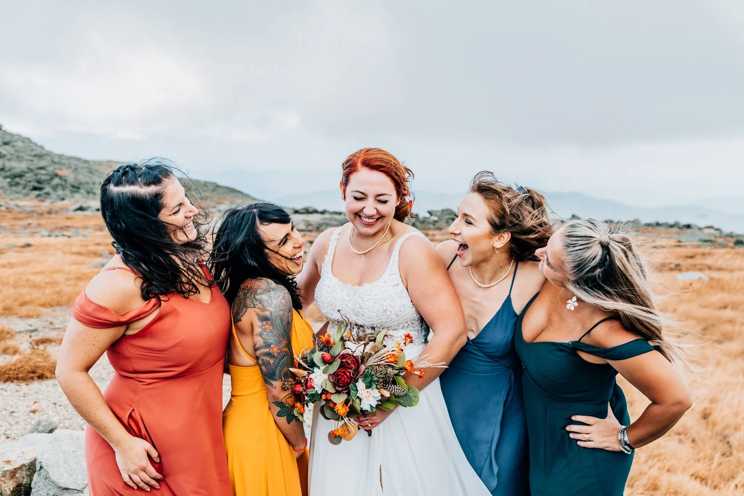 Bride laughing with four bridesmaids in rust, marigold and navy dresses on the Mount Washington plateau