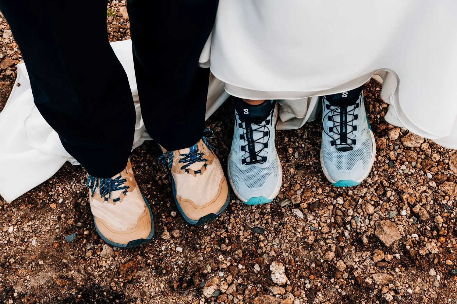 Detail of trail running shoes worn beneath a wedding dress and suit on a mountain summit