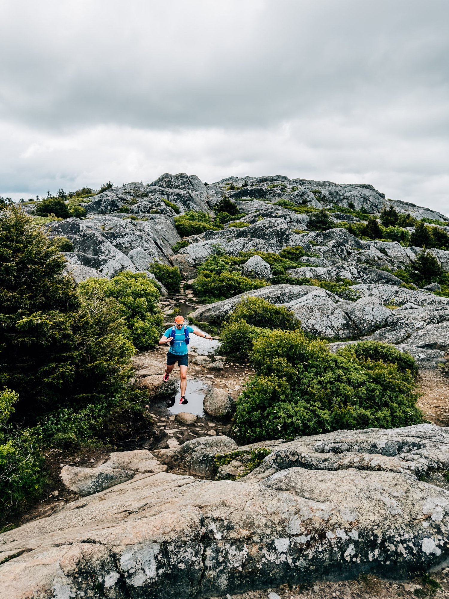 Trail runner in a blue shirt and orange cap hopping across a puddle on the granite summit of Mount Monadnock under a heavy gray sky