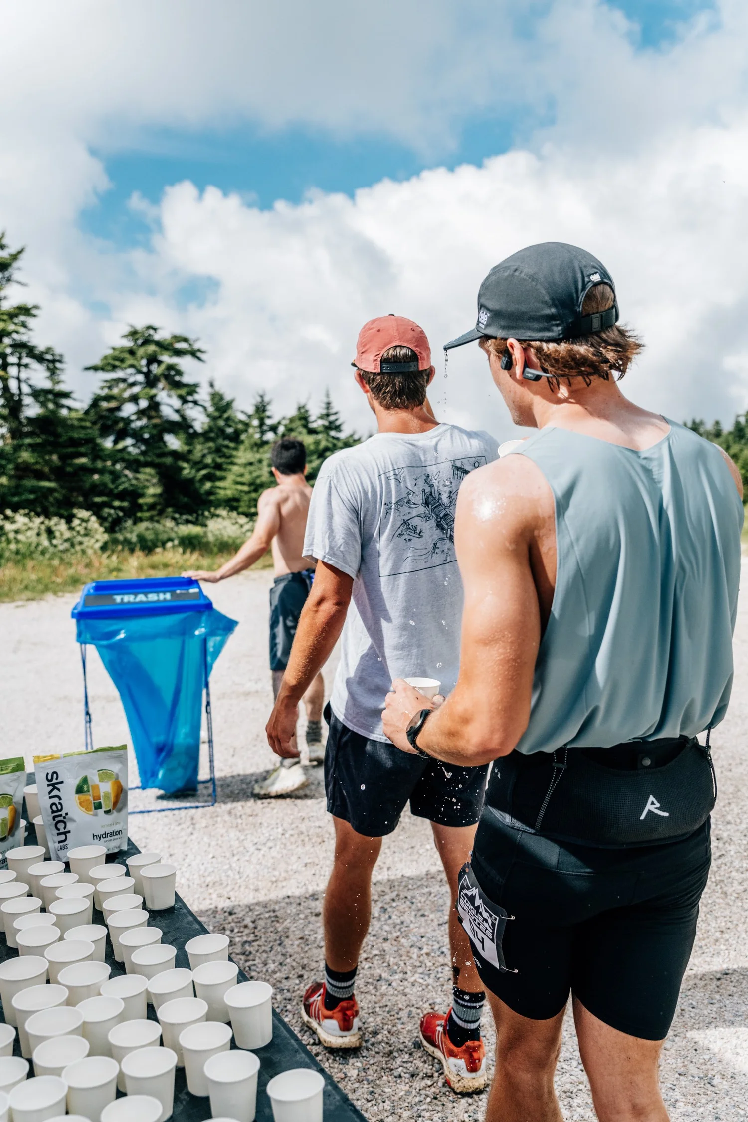 Runners grabbing paper cups of water at a summit aid station table under big clouds