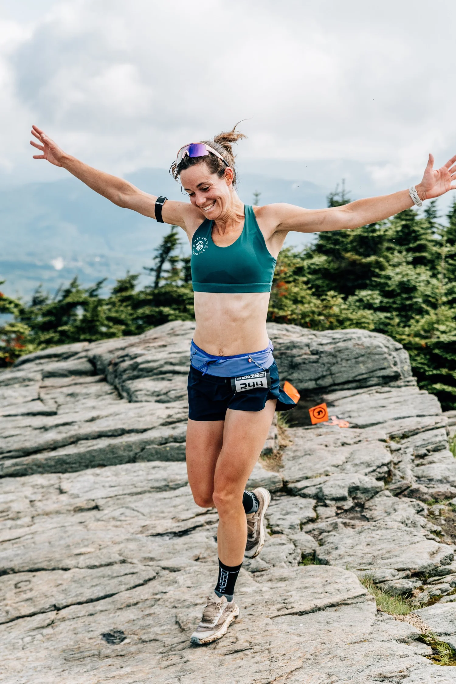 Smiling runner with arms outstretched hopping across granite ledges on the course, a Cirque Series bib at her waist