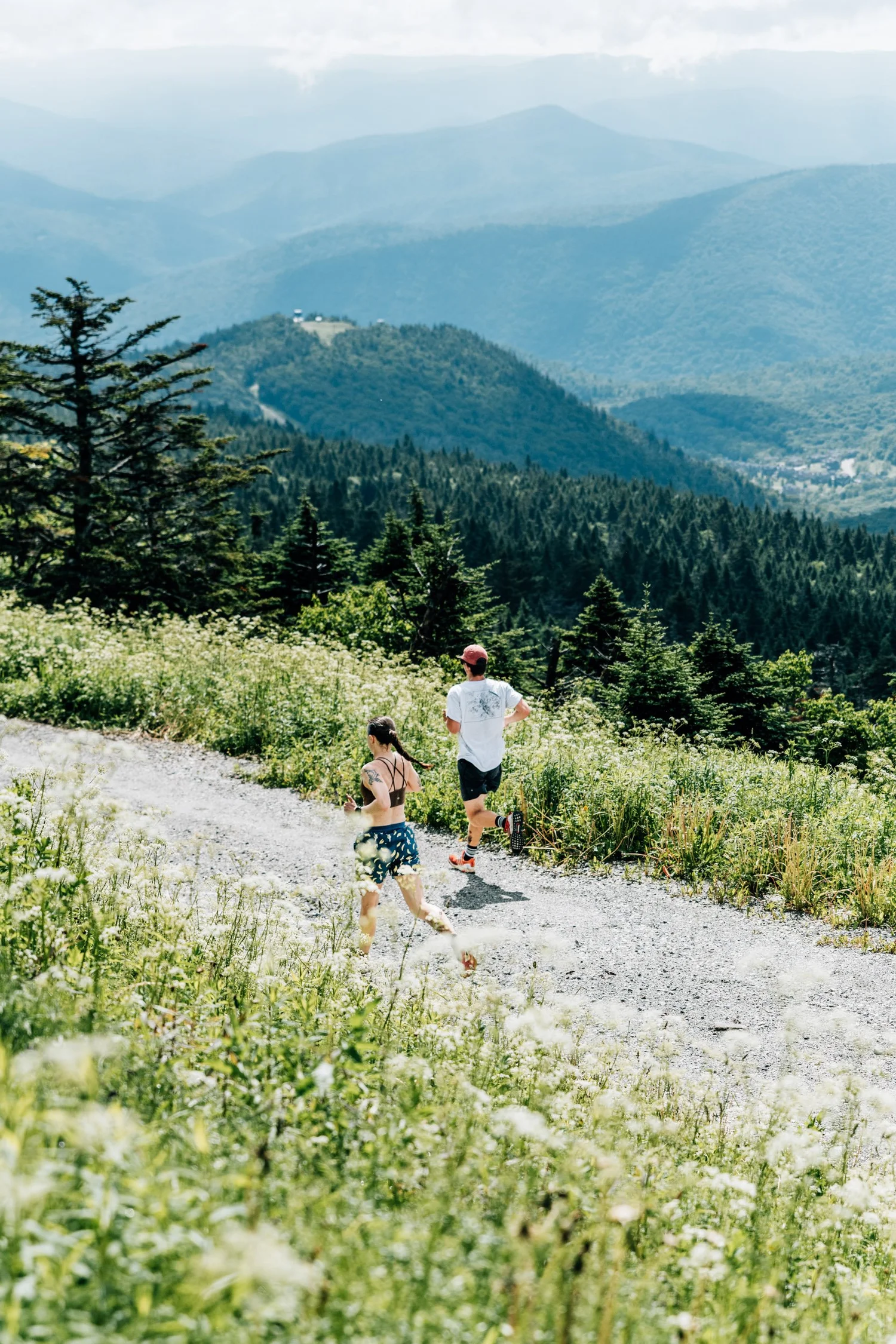 Two runners heading down a gravel road through white wildflowers with layered green mountains behind them