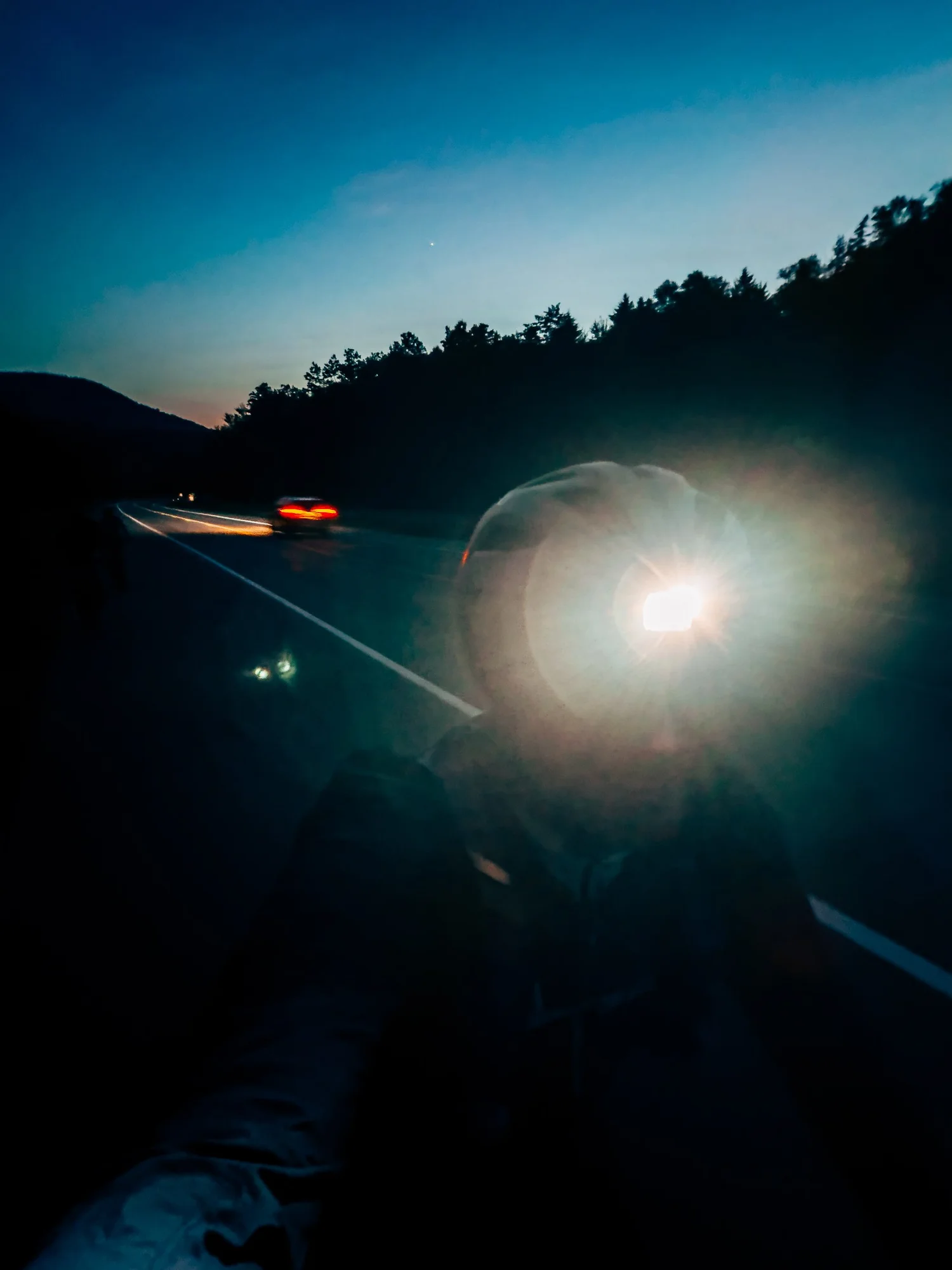 Cyclist with a bright headlight beside a darkening road at dusk as a car&rsquo;s taillights glow in the distance