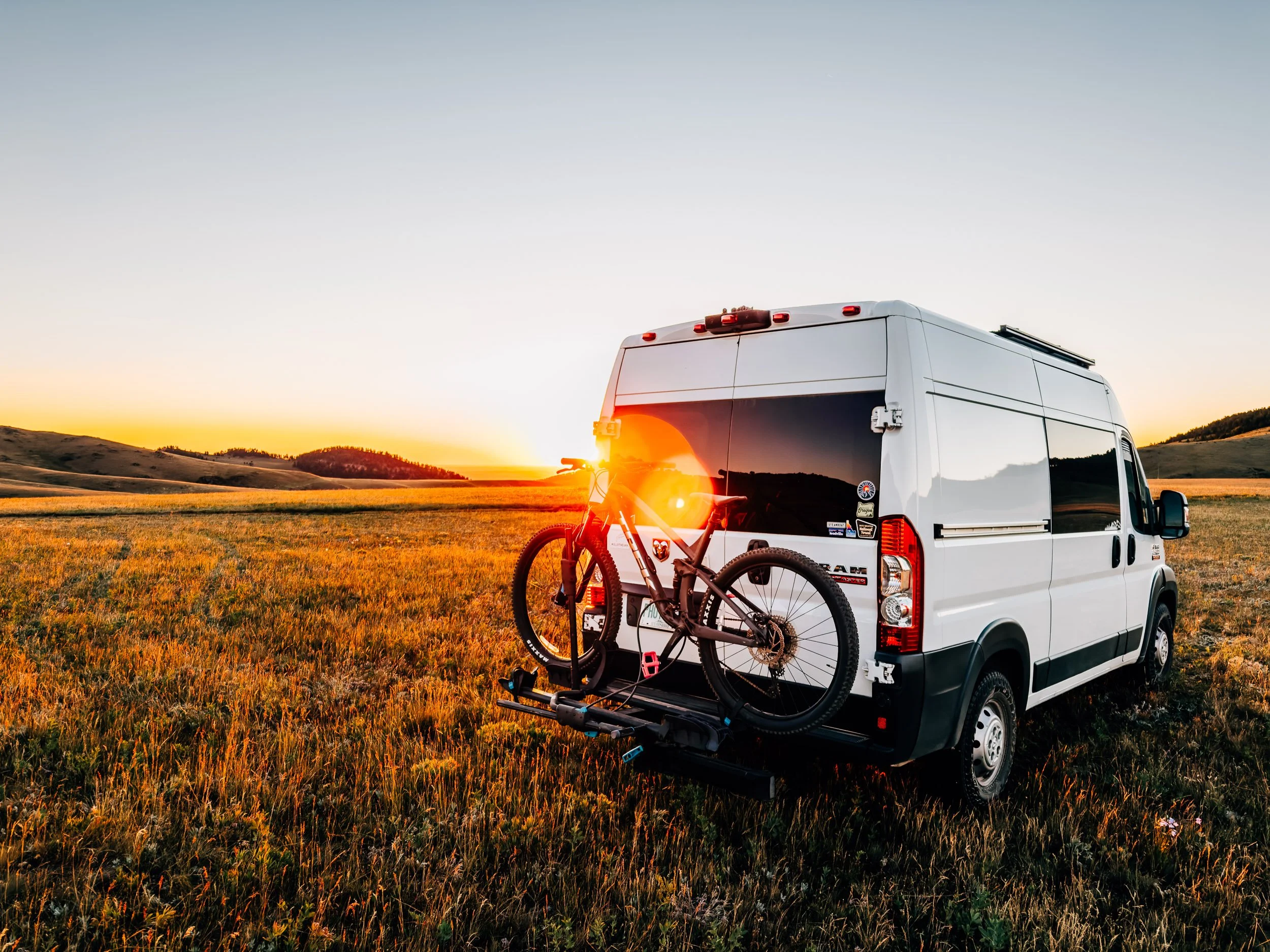 White ProMaster van with a mountain bike on the rear rack parked in a golden grass meadow as the sun sets over rolling hills