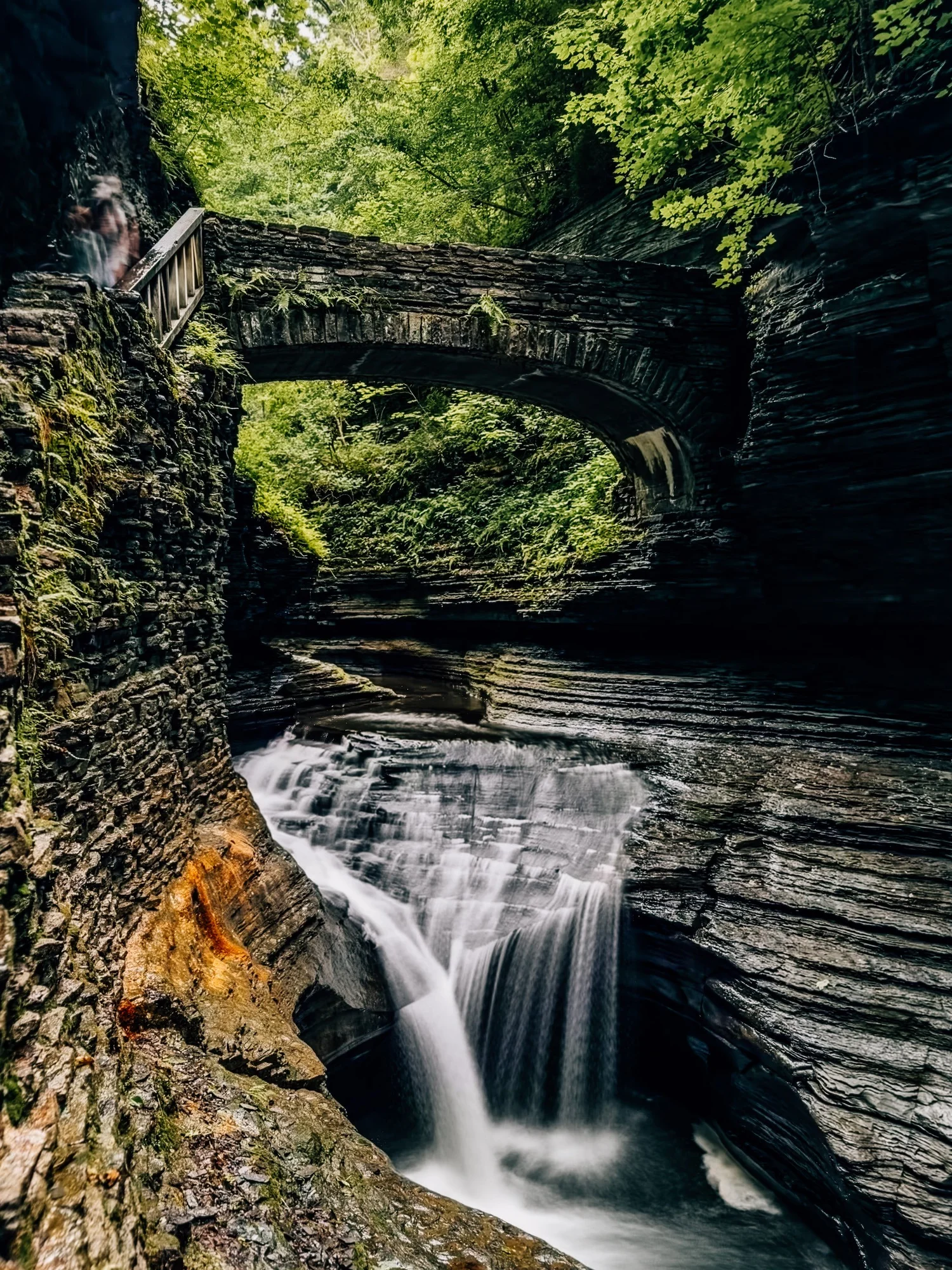 Stone bridge arching over a narrow, mossy gorge with a waterfall dropping into a dark pool below
