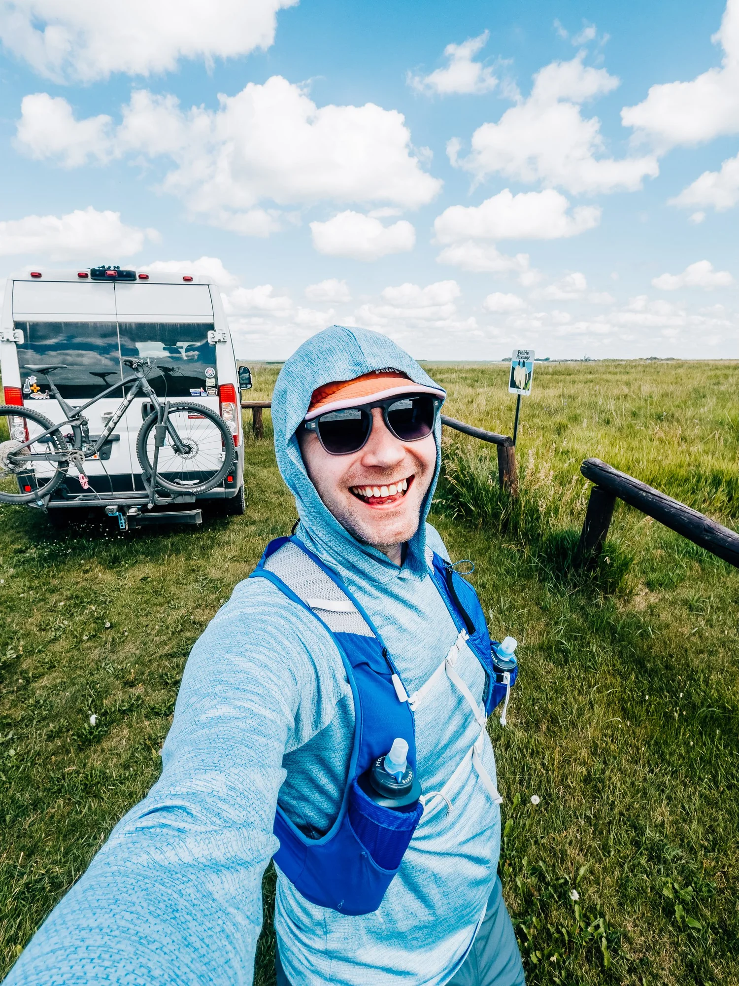 Smiling runner in a blue hoodie and running vest taking a selfie on a green prairie, the van and bike rack behind him