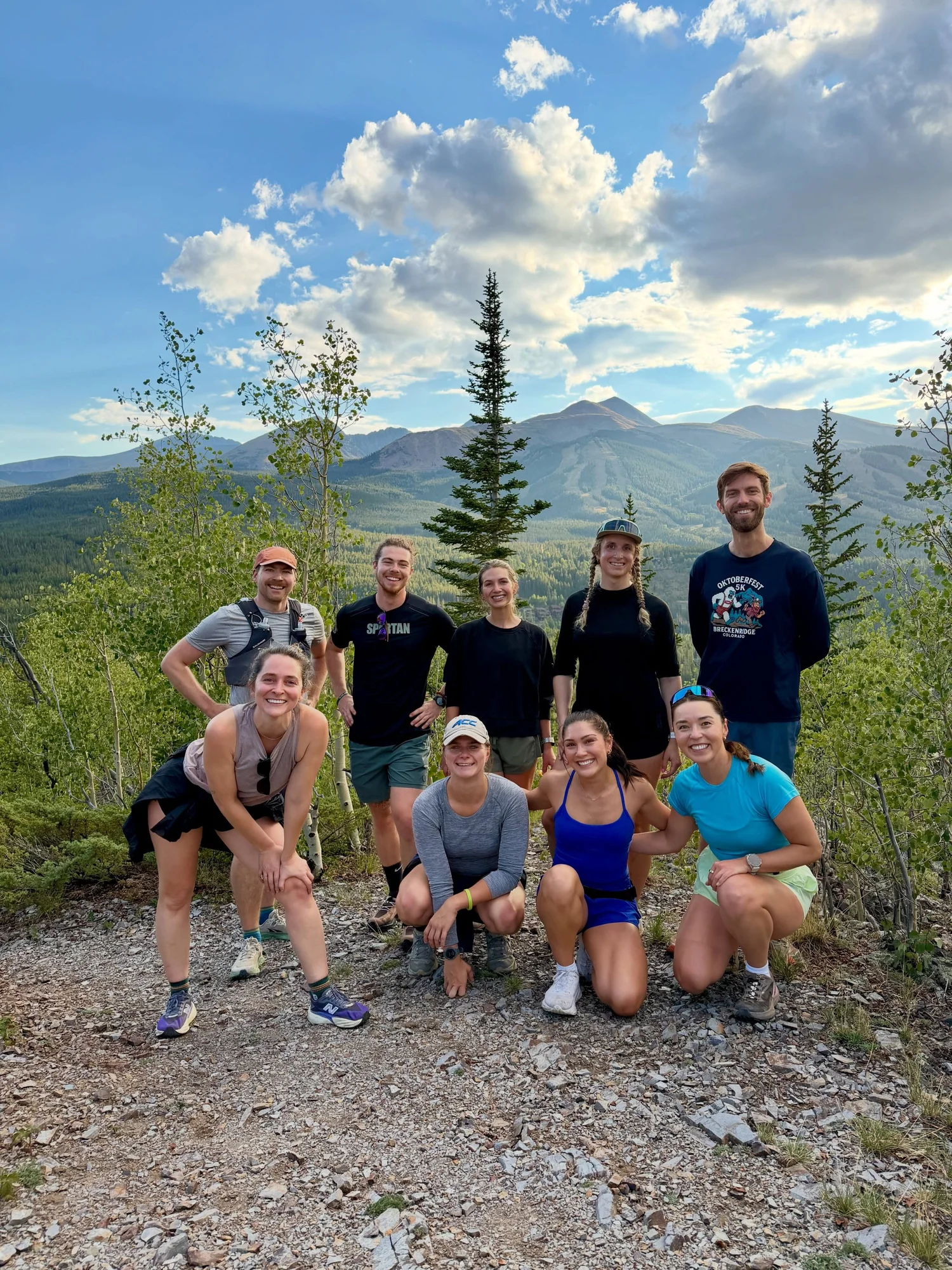 Nine smiling runners in two rows on a gravel overlook with aspen, spruce and a wide mountain valley behind them
