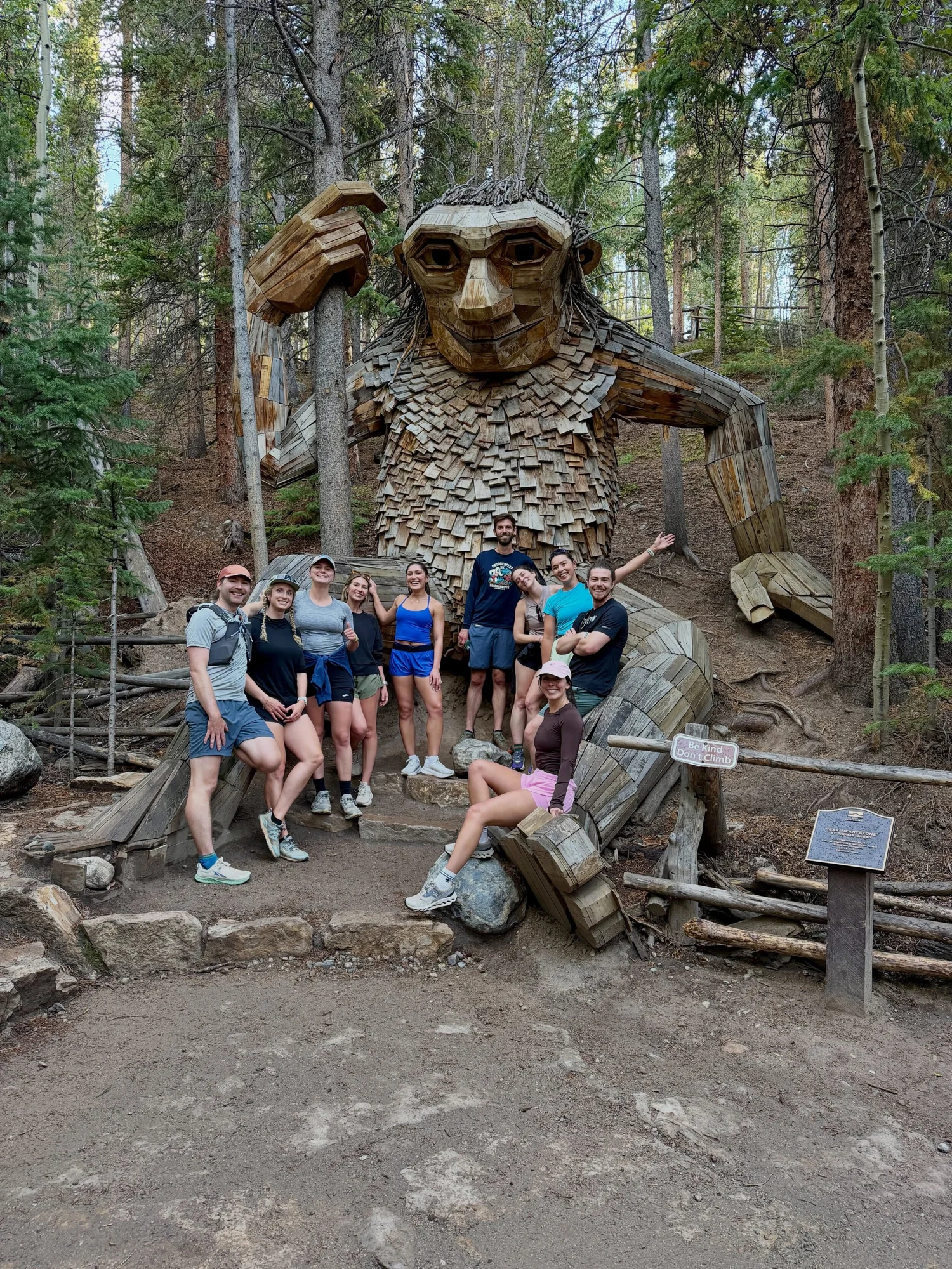 Group of ten runners posing around a giant wooden troll sculpture in a pine forest, a sign reading Be Kind Don&rsquo;t Climb beside them