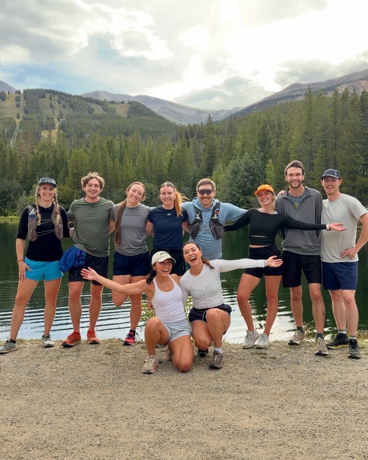 Group of runners laughing with arms around each other in front of a calm mountain lake and forested hillside
