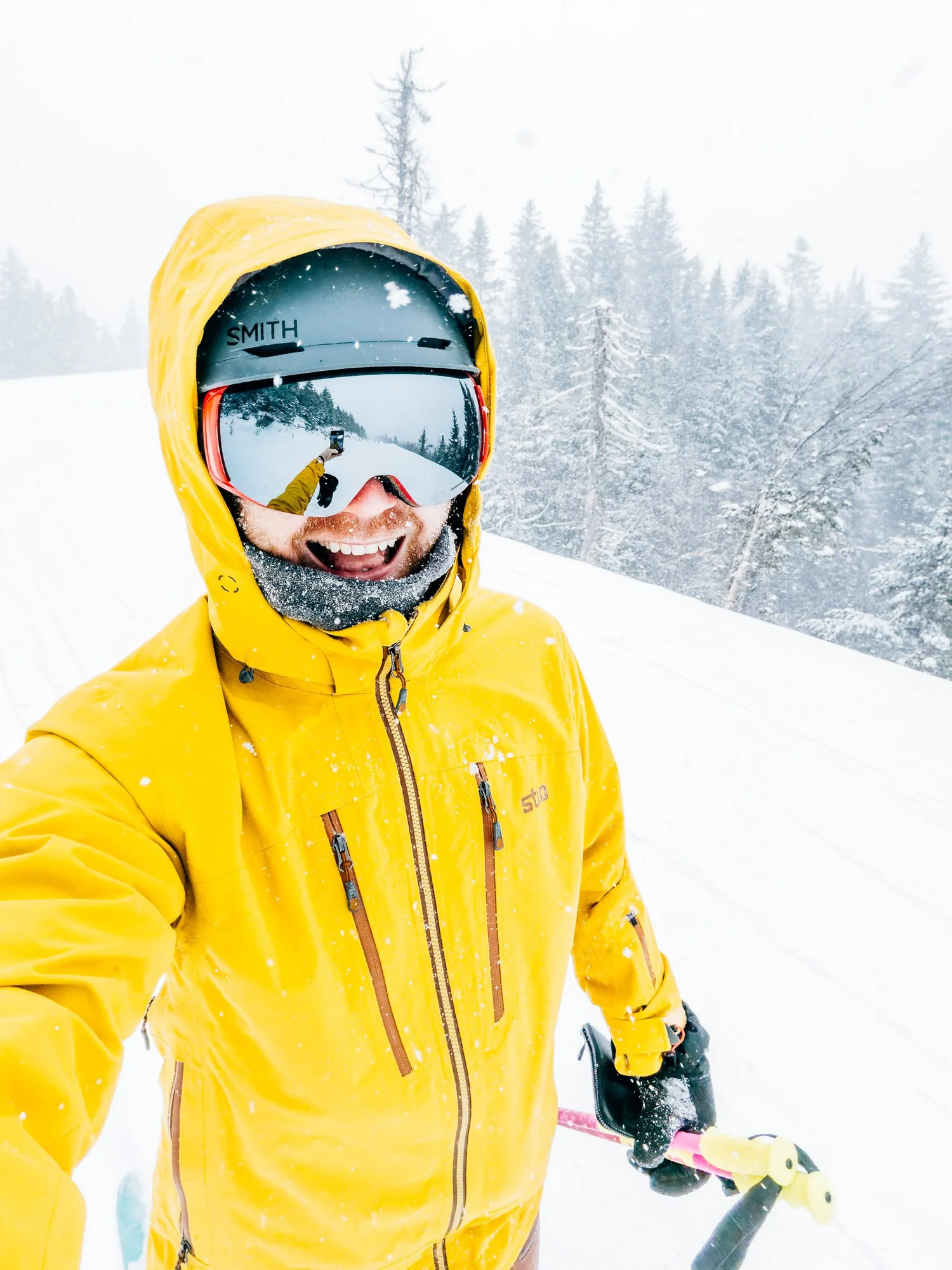 Skier in a yellow jacket, helmet and mirrored goggles laughing in a selfie as snow falls on a groomed run